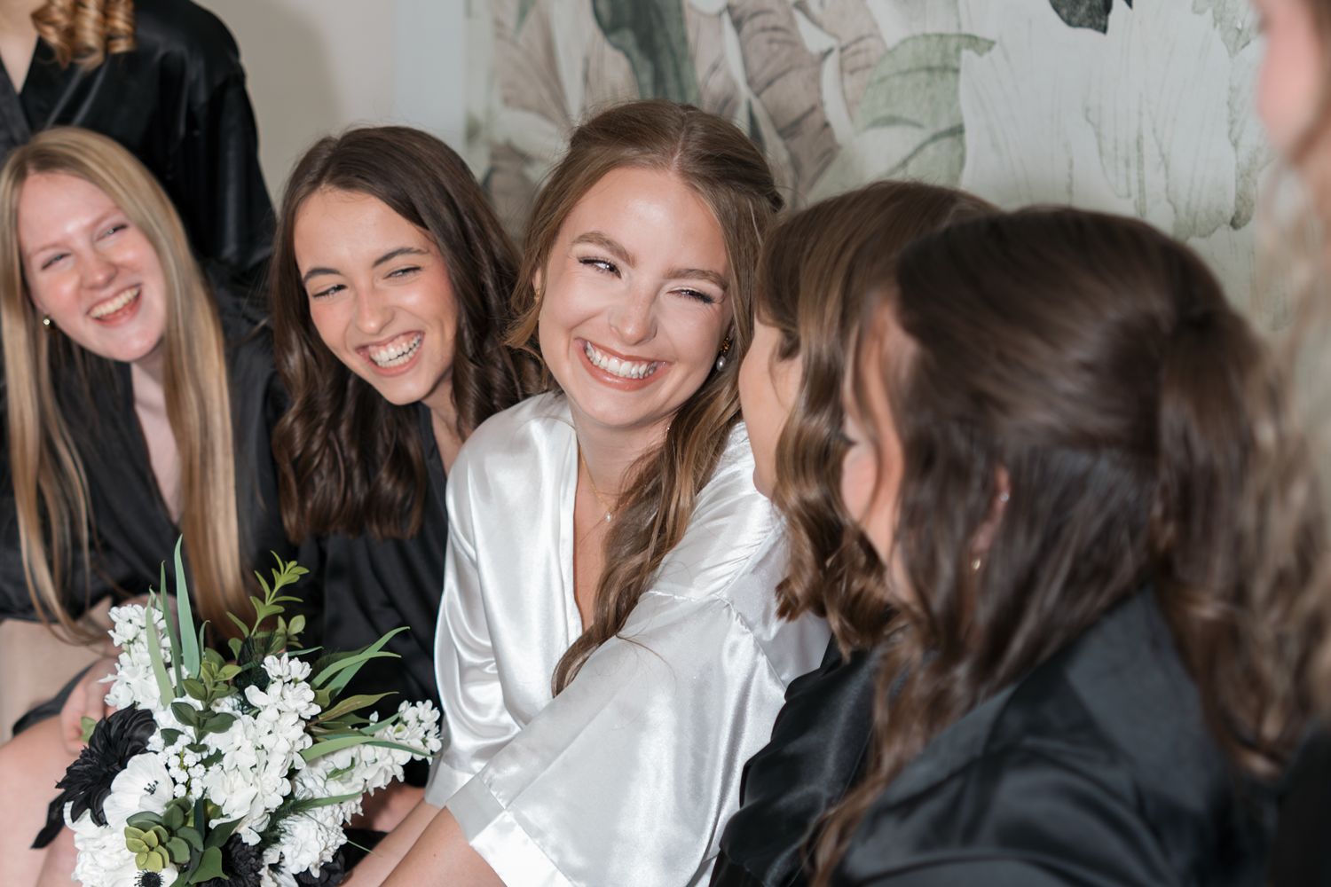 Bride in white satin robe laughing with bridesmaids while holding white floral bouquet during wedding preparations.