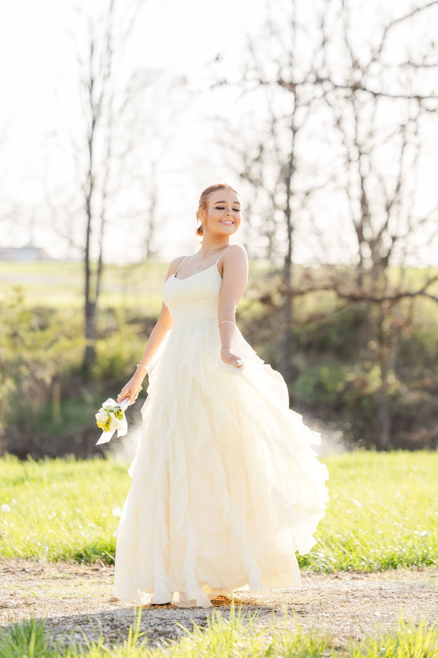 A bride in a flowing ivory gown twirls gracefully in a sun-drenched field with bare trees in the background.