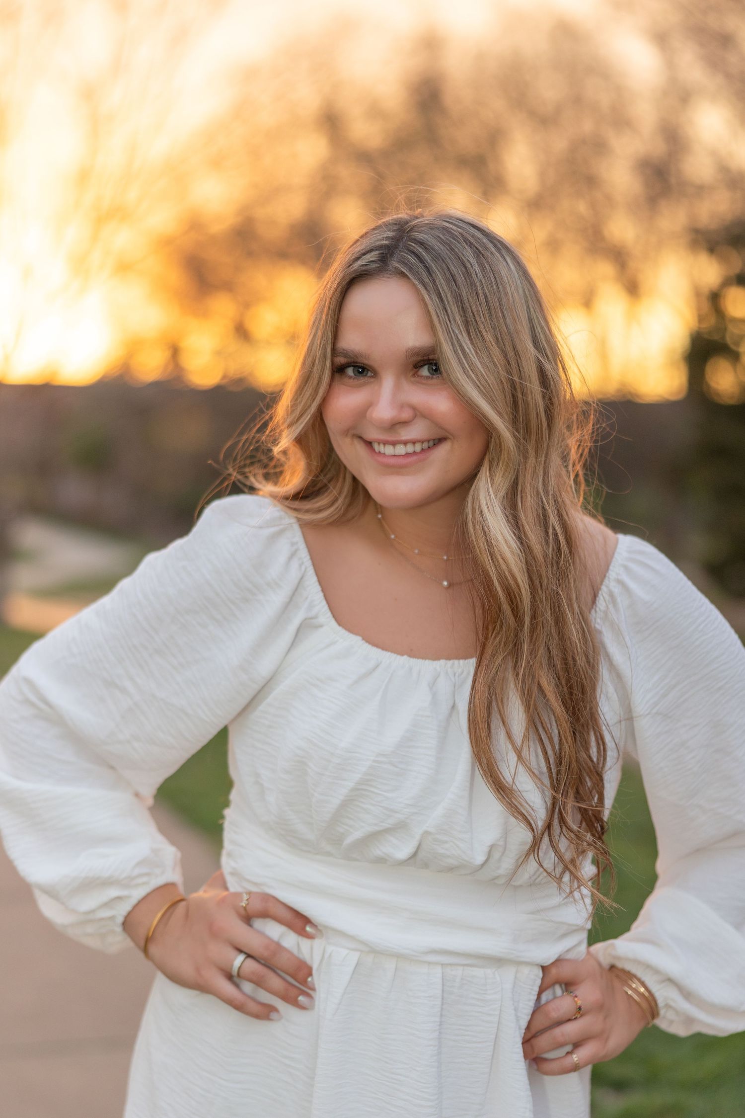 Portrait of someone in a flowing white dress with long blonde hair smiling outdoors during golden hour sunset.