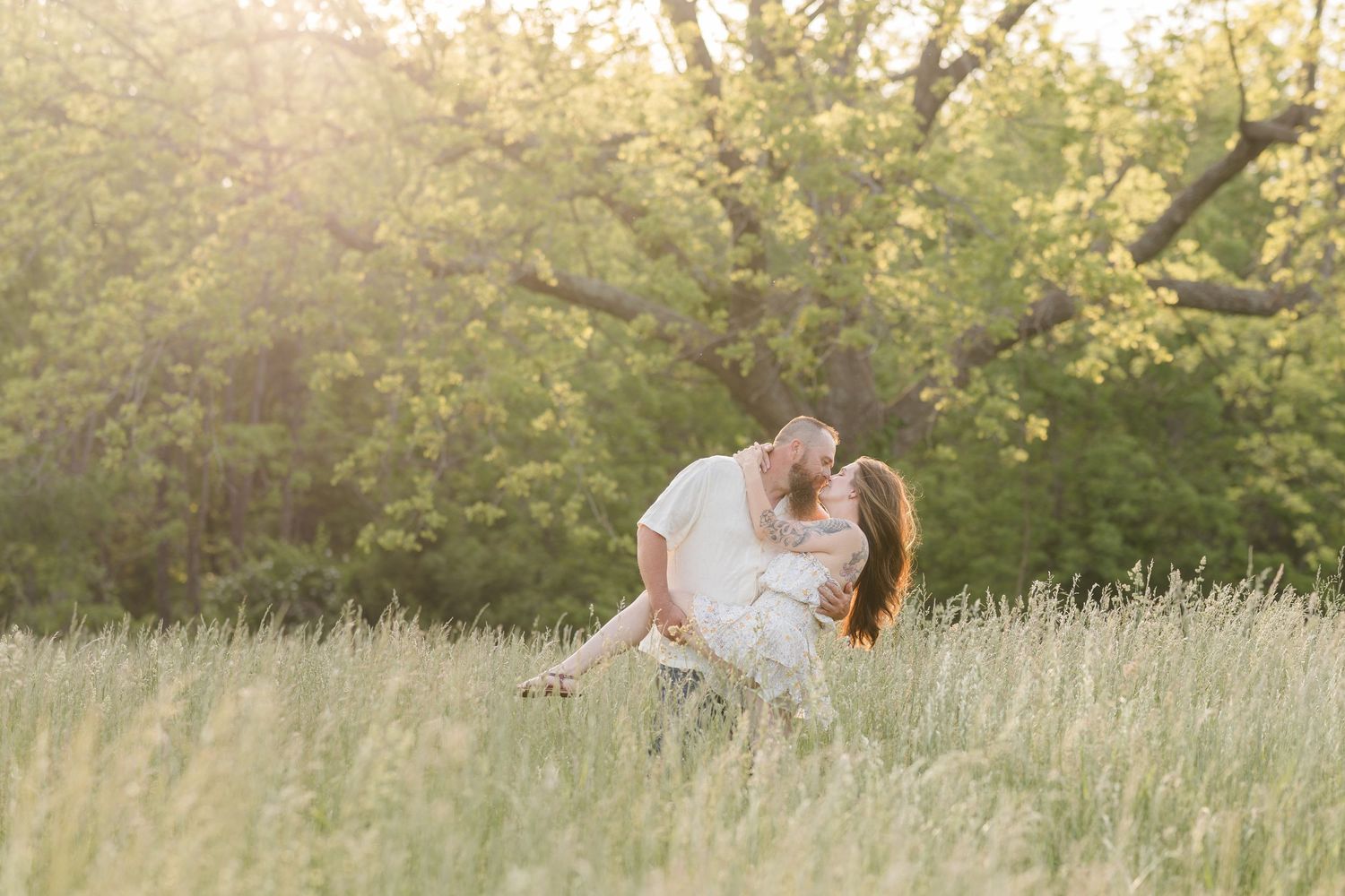 A couple in white embraces in a sunlit meadow with golden grasses and blooming trees in the background.