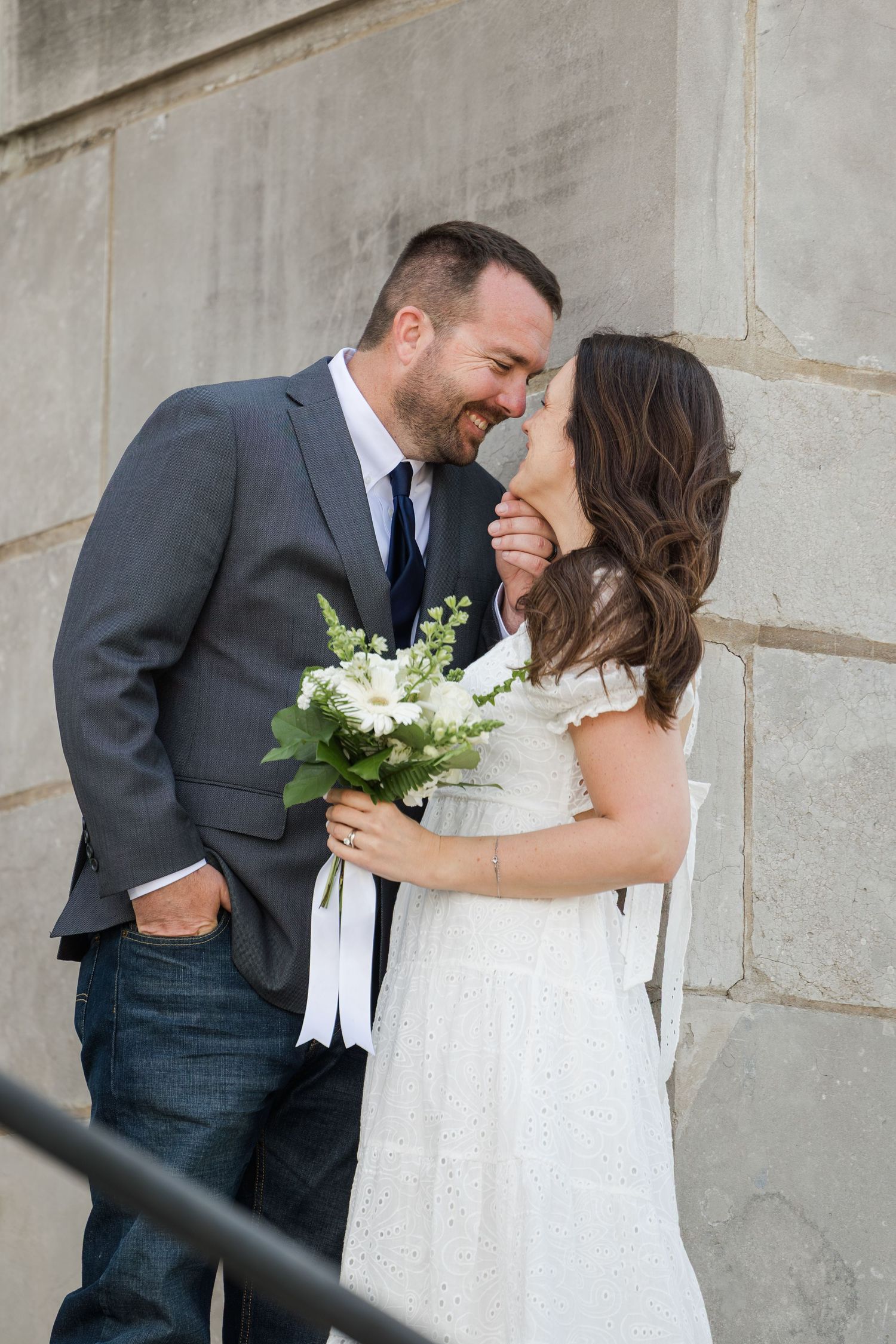 A romantic wedding moment with a couple embracing against a stone wall, the bride holding a white floral bouquet.