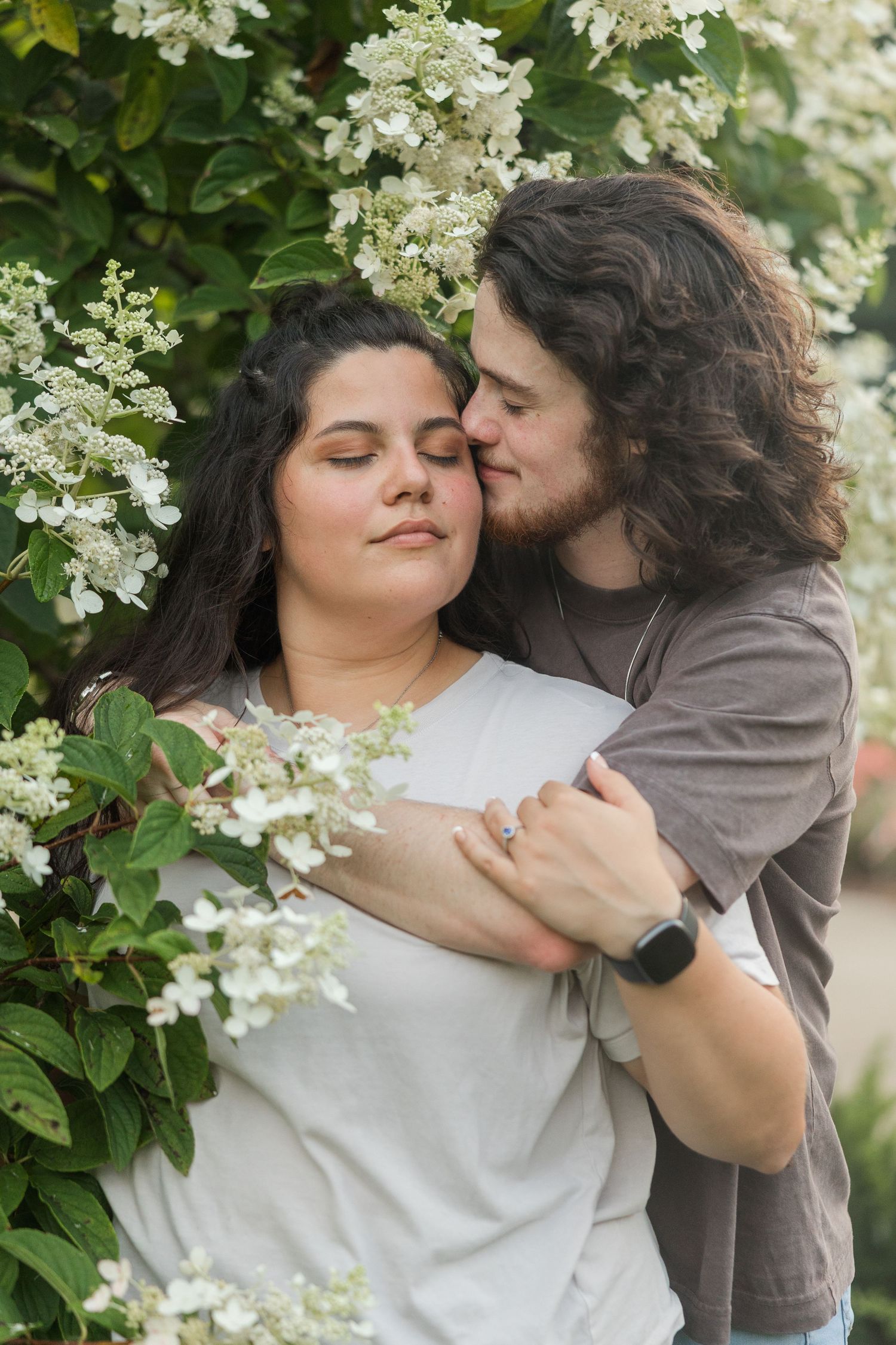 Romantic couple embraces among blooming white hydrangea flowers in garden setting.
