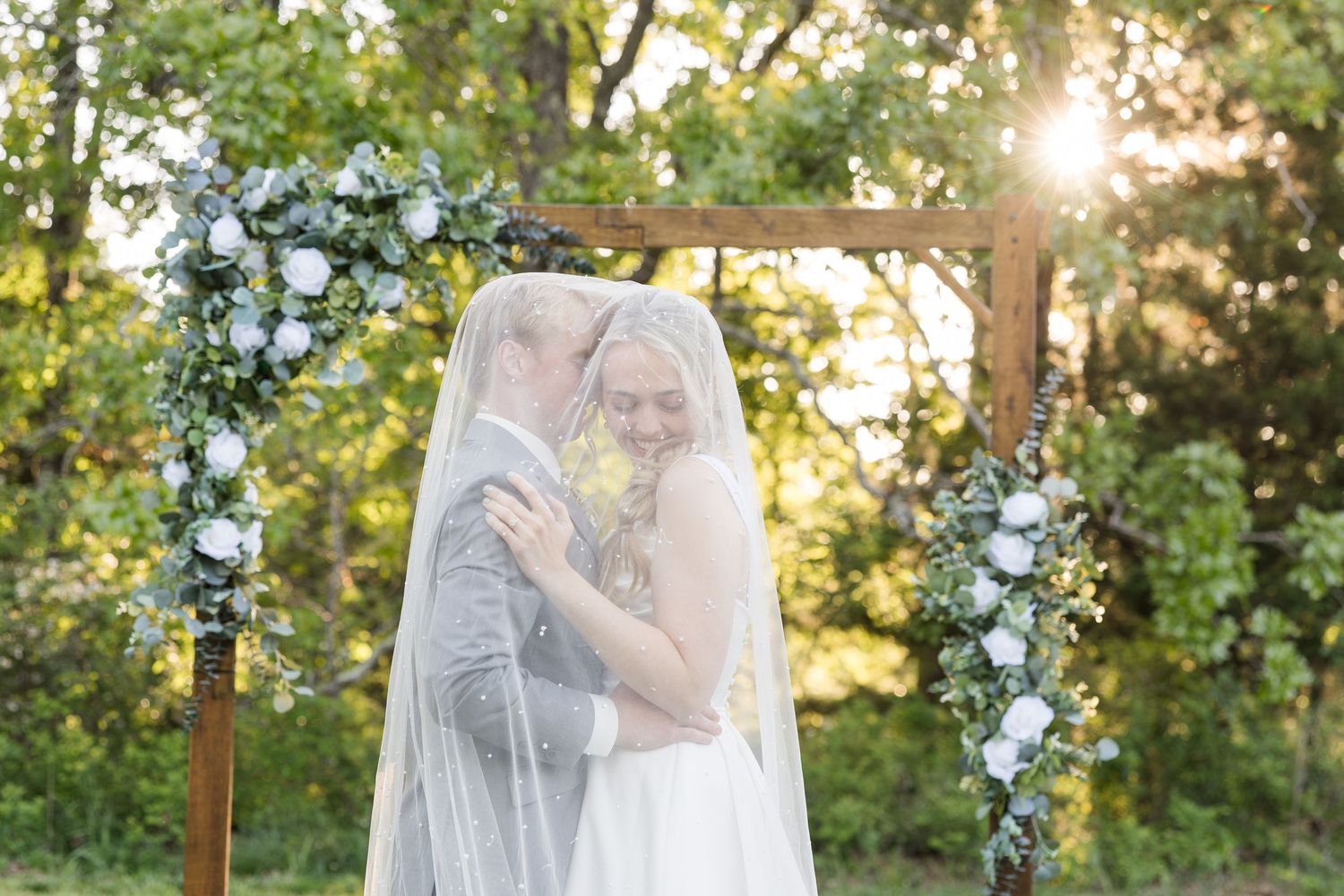 Bride and groom share a romantic moment under a floral wooden arch outdoors, draped in a flowing white veil.
