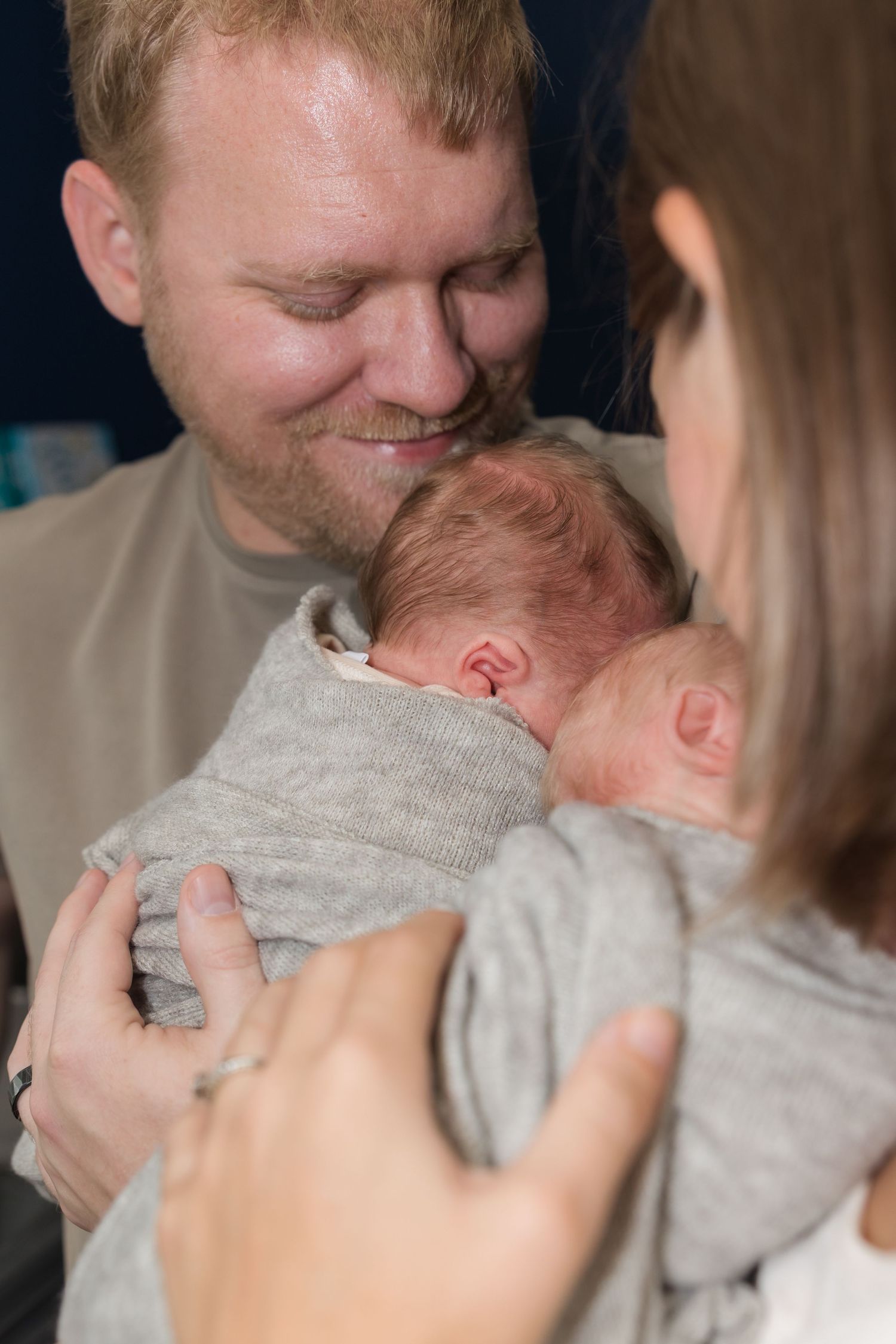 Happy father and mother lovingly cradling their newborn baby wrapped in a gray swaddle blanket.