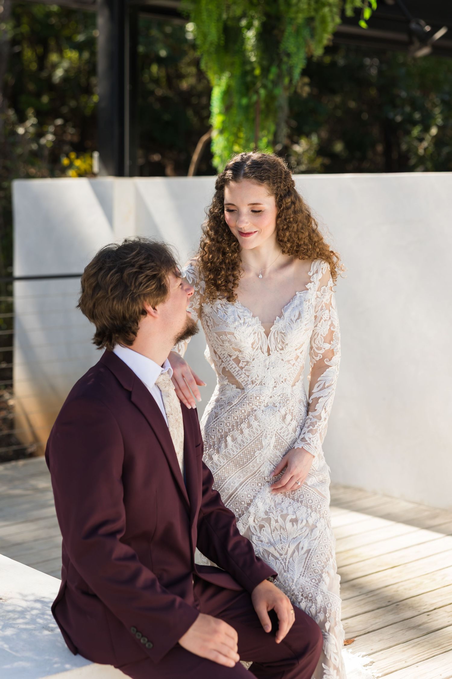 Bride in lace gown smiles at groom kneeling before her on sunny outdoor wedding venue deck.