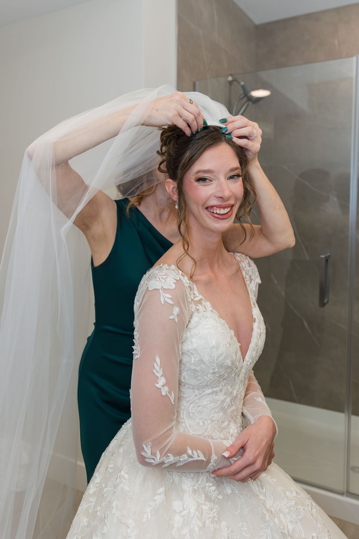 Smiling bride in lace wedding gown gets veil placed by bridesmaid in green dress during wedding preparations.