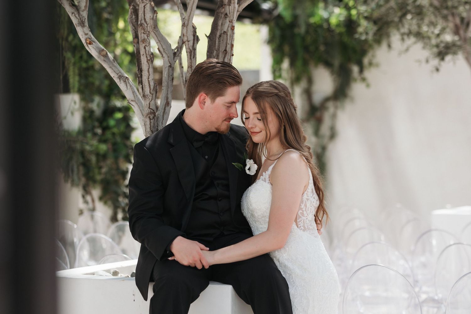 Bride and groom share a romantic moment in elegant wedding attire, surrounded by lush greenery at an outdoor venue.