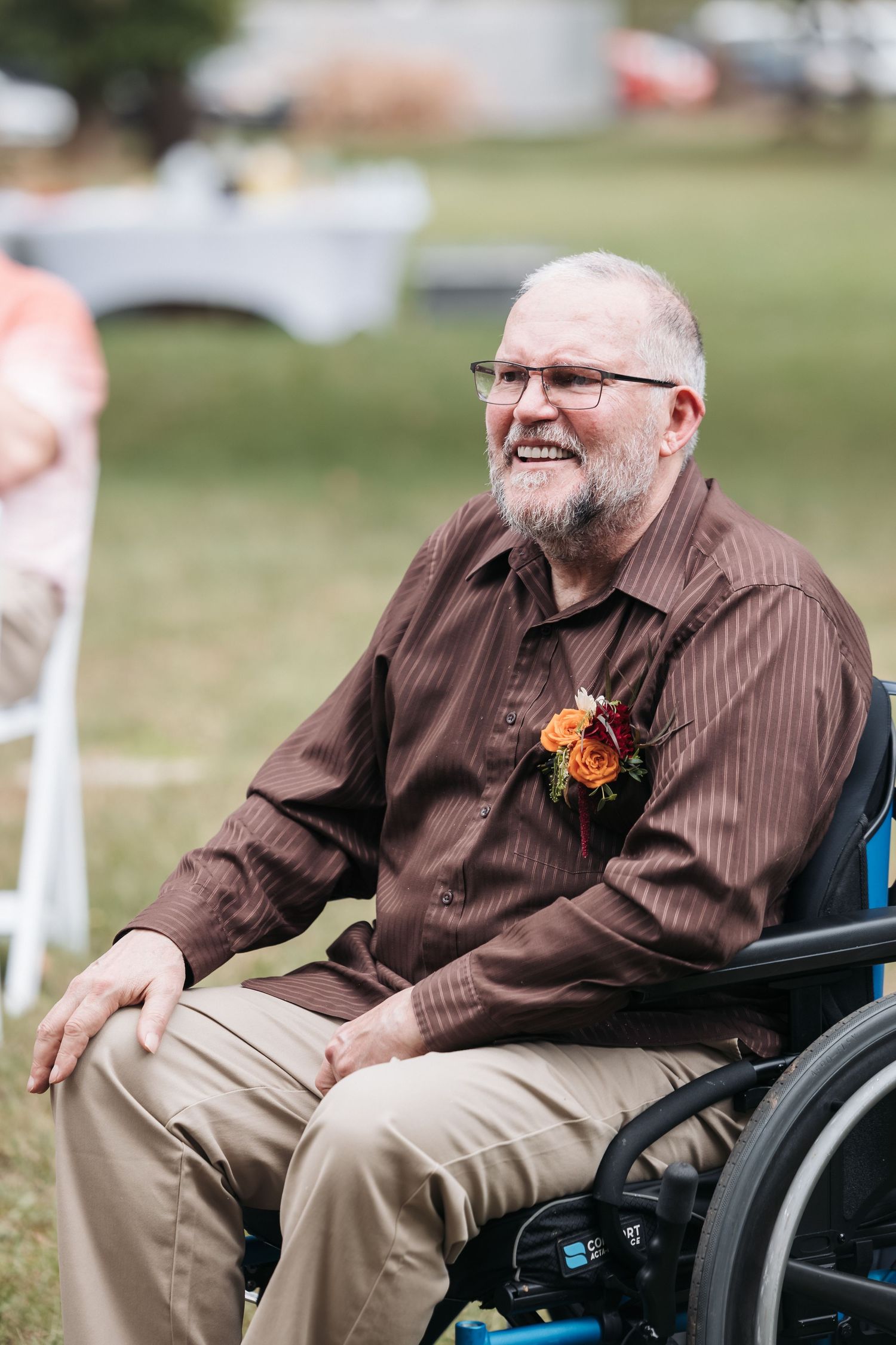 Smiling elderly man in wheelchair wearing brown shirt with floral boutonniere at outdoor wedding ceremony.