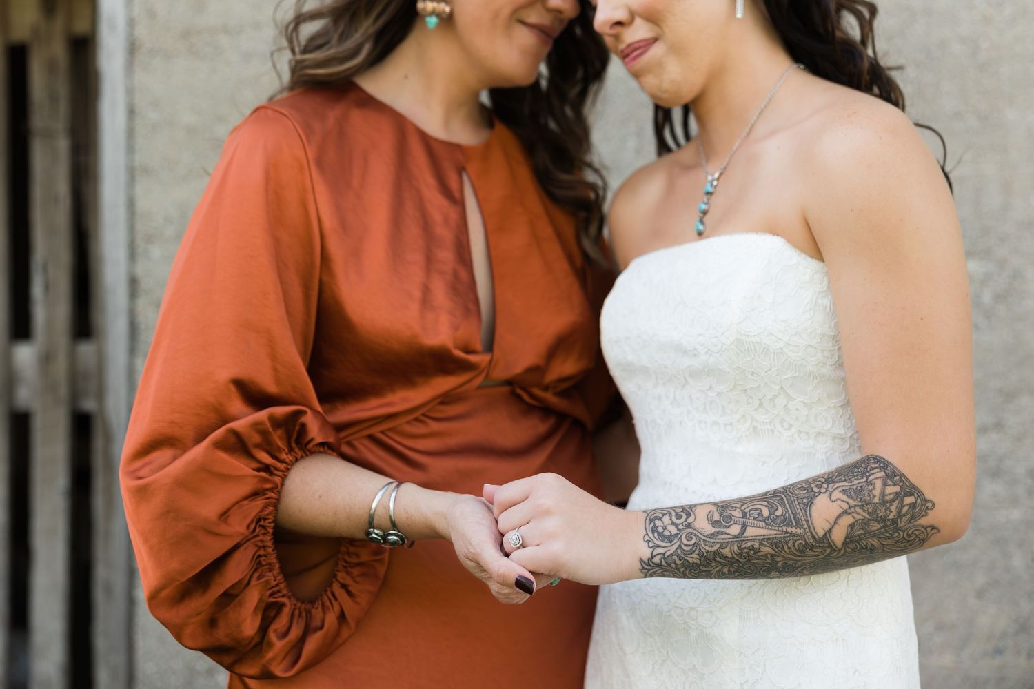 Bride in white strapless gown with sleeve tattoo holds hands with woman in rust orange dress at wedding.