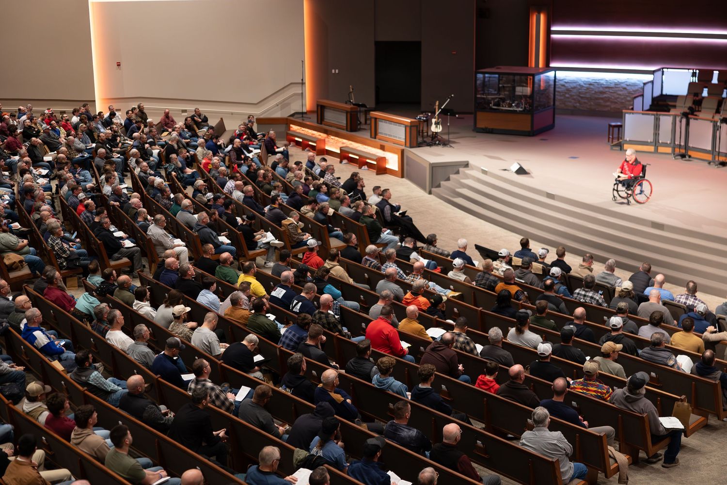 A large audience fills the seats of an auditorium during a presentation on stage with red lighting.