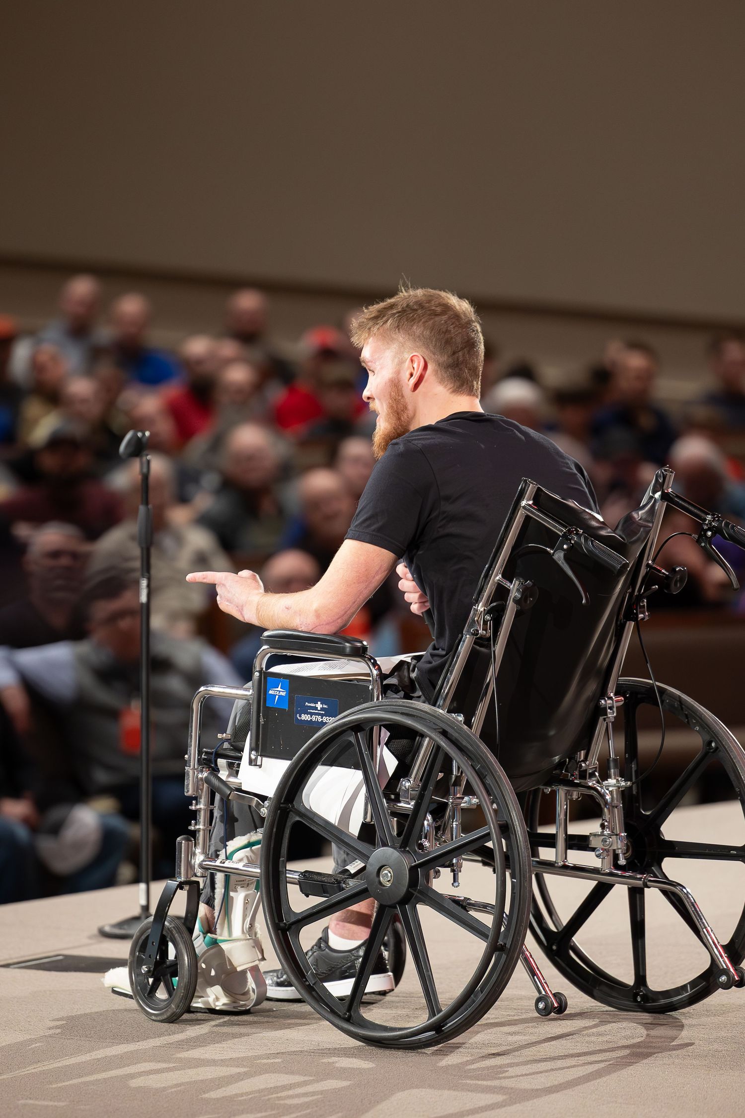 A speaker in a wheelchair addresses an audience from a stage during a presentation.