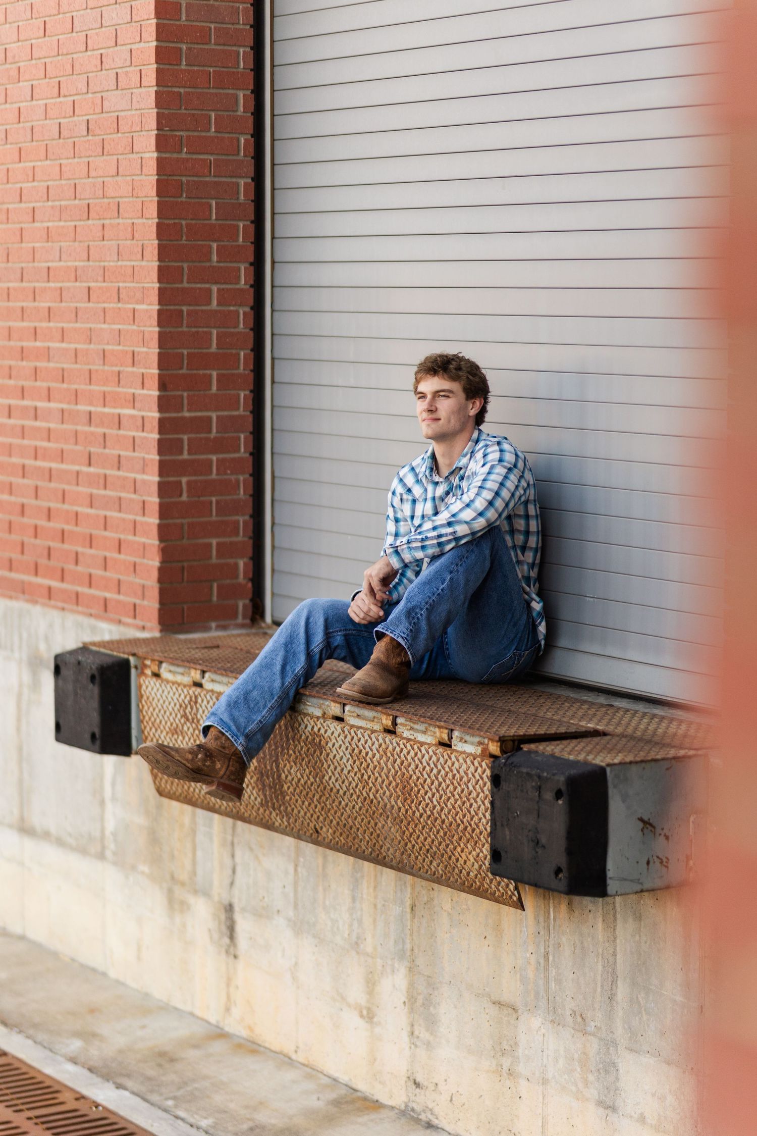 A casual outdoor portrait of someone sitting on a loading dock wearing a plaid shirt and jeans.