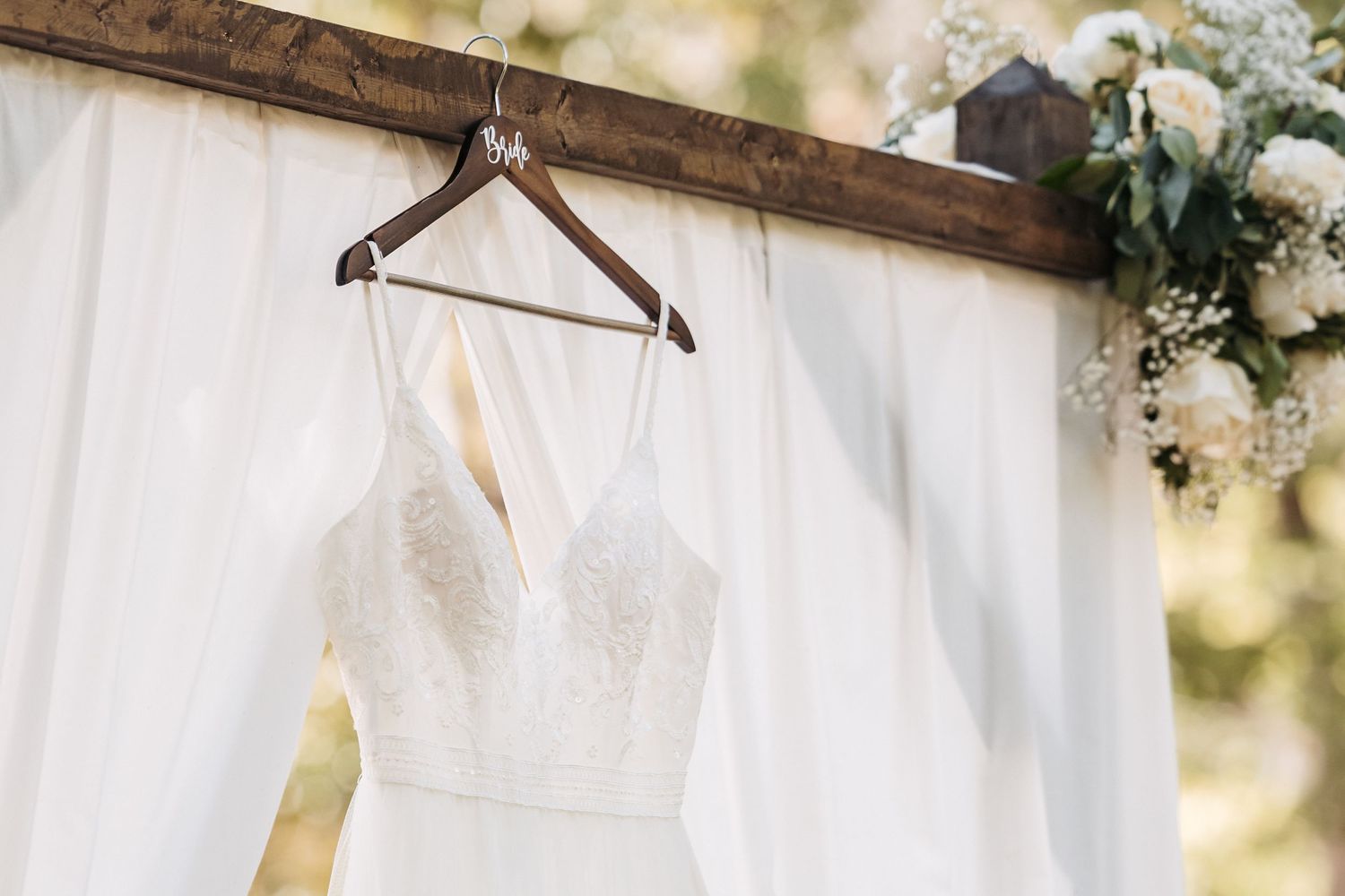 White wedding dress on wooden hanger against white drapery with floral decorations.