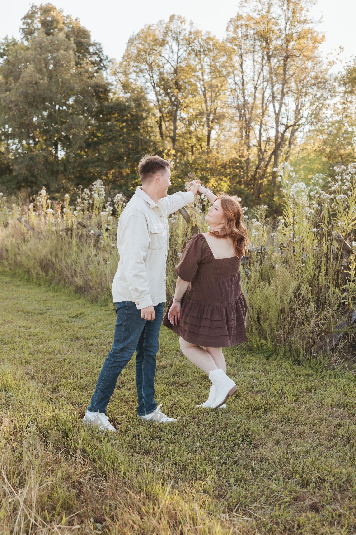Couple dancing playfully in a field at golden hour wearing white and brown outfits.