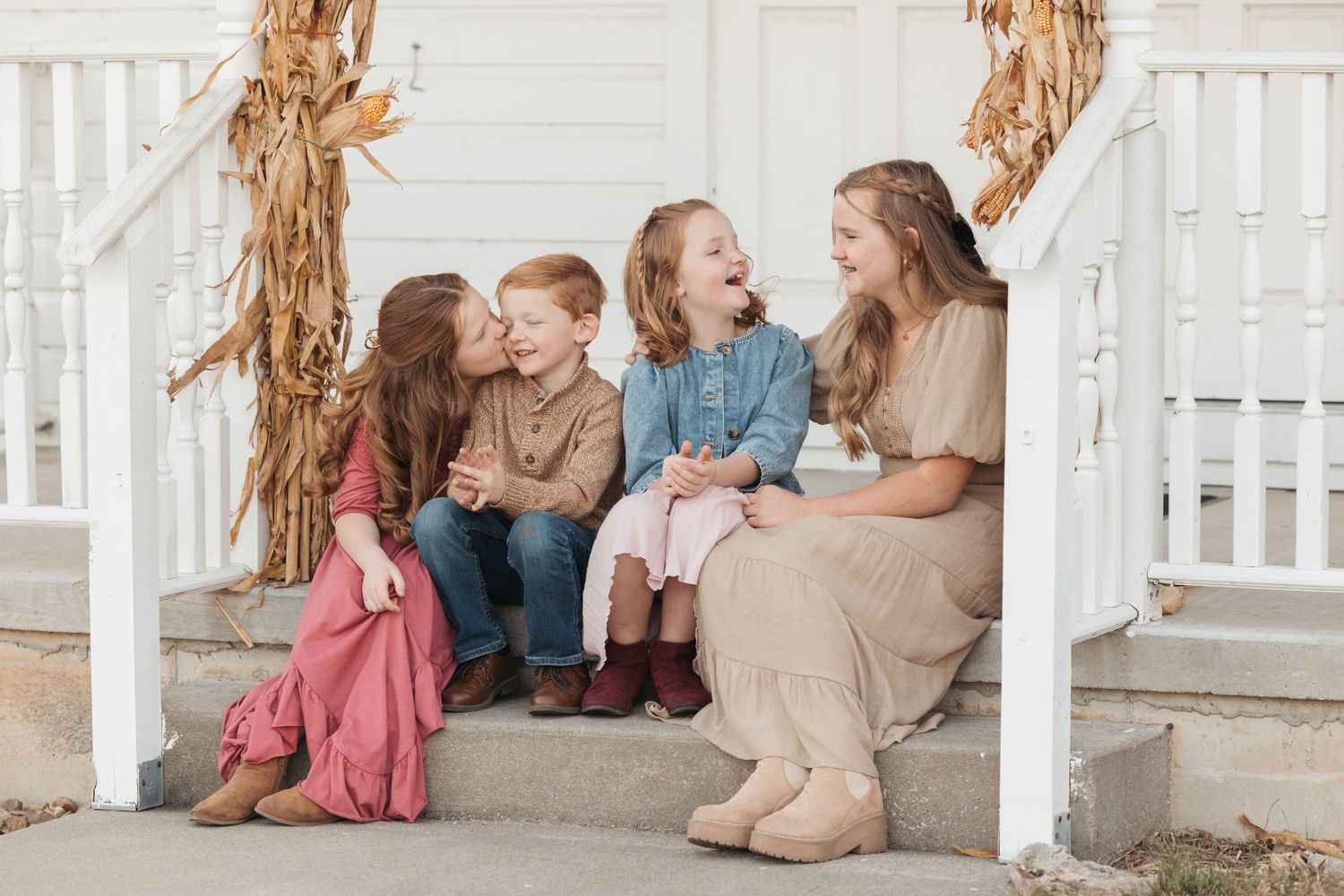 Family sitting together on white porch steps decorated with autumn corn stalks.