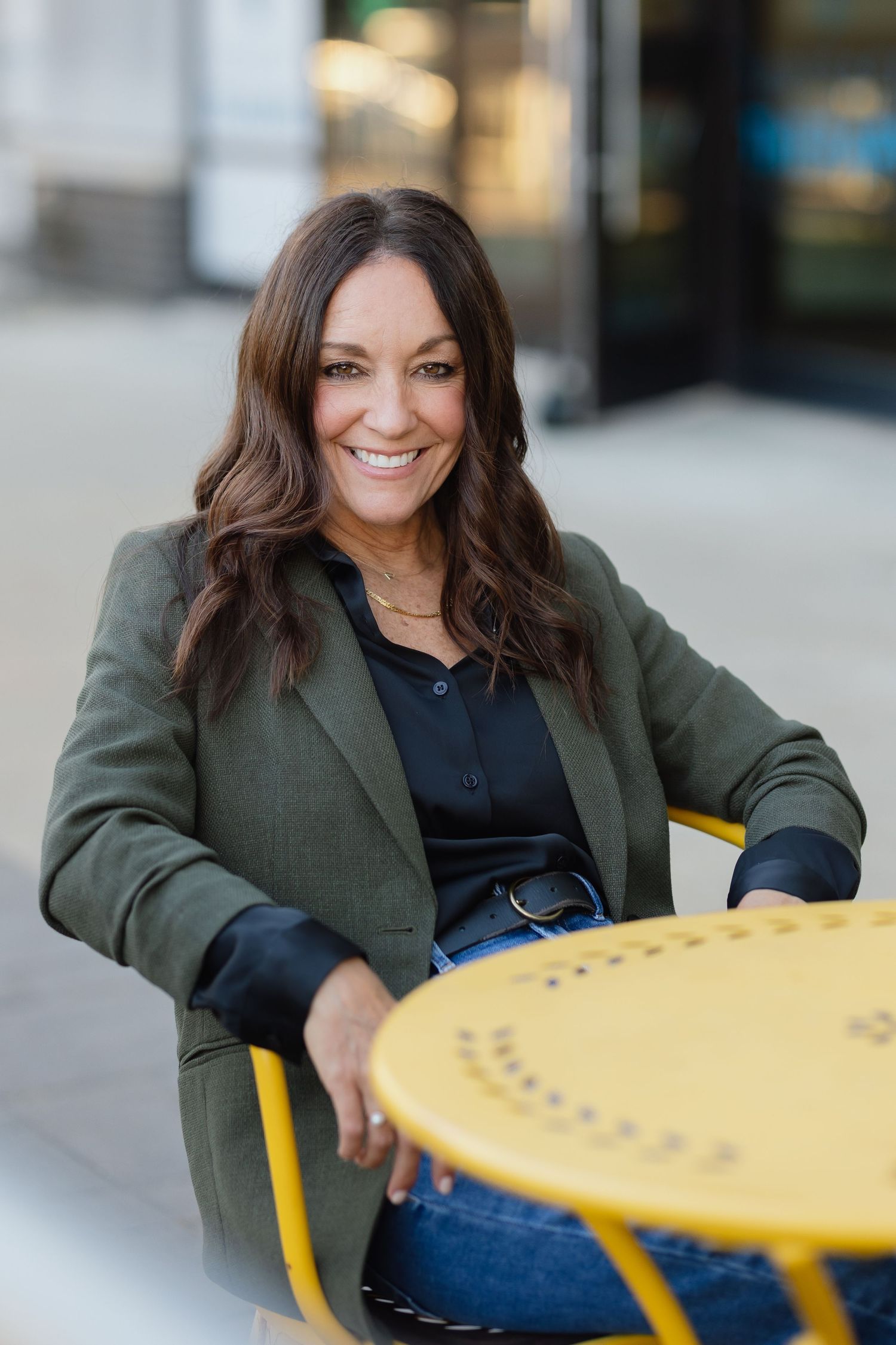 Professional portrait of a woman in green blazer seated at yellow outdoor table.