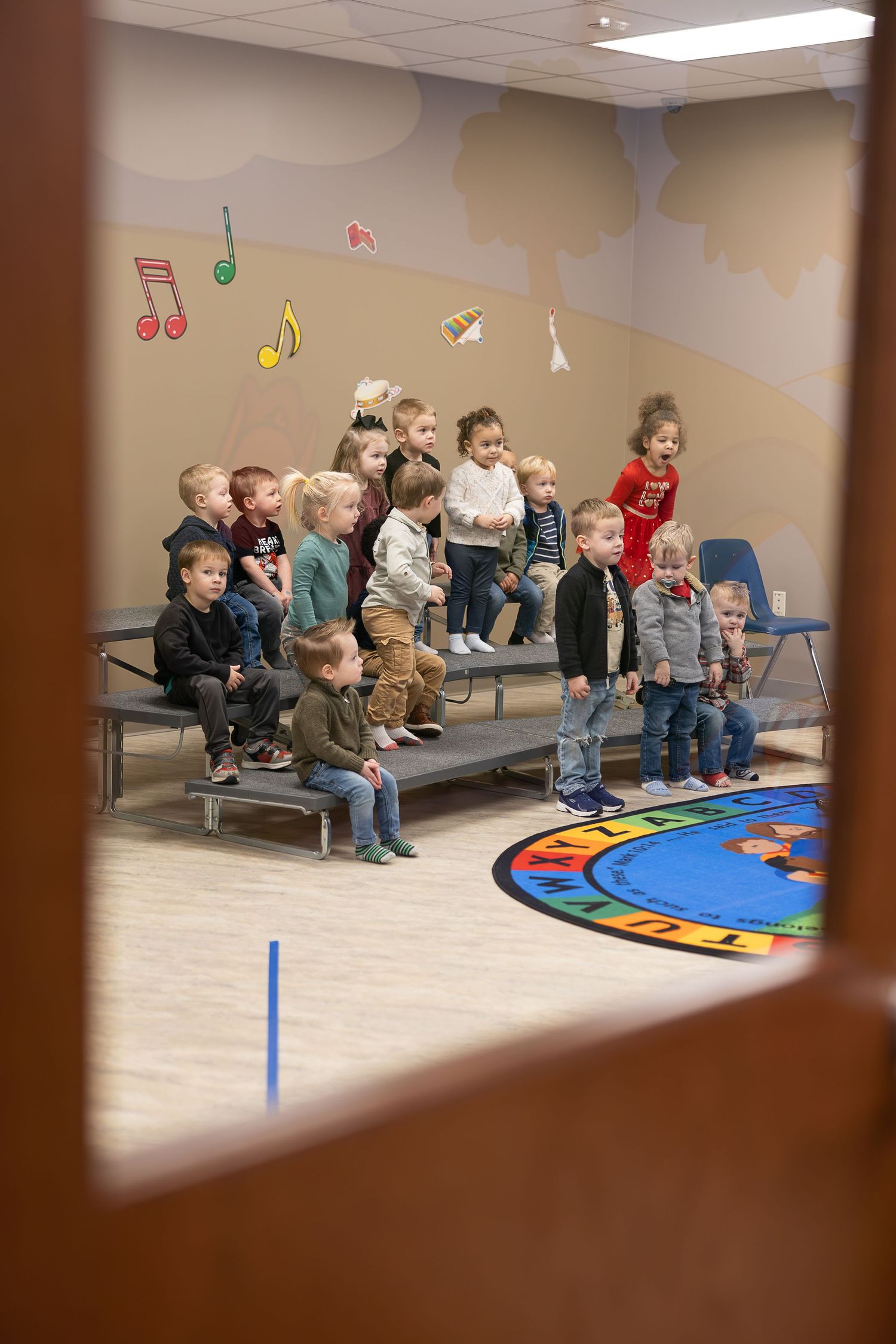 Young students sit on risers in a classroom with musical note decorations on the wall.