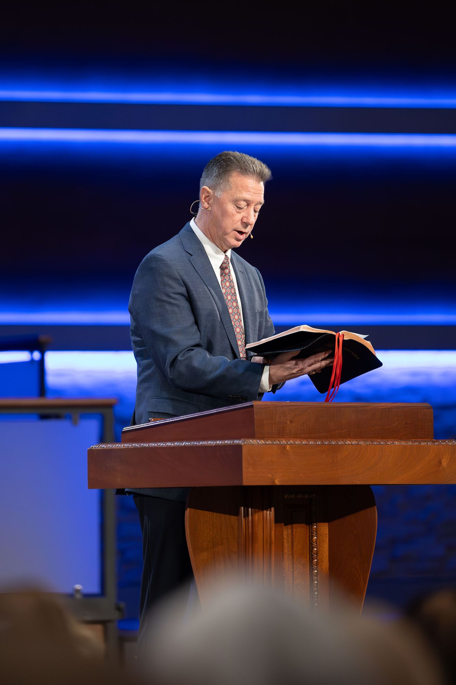 Speaker stands at wooden podium reading from Bible with blue lighting in background.