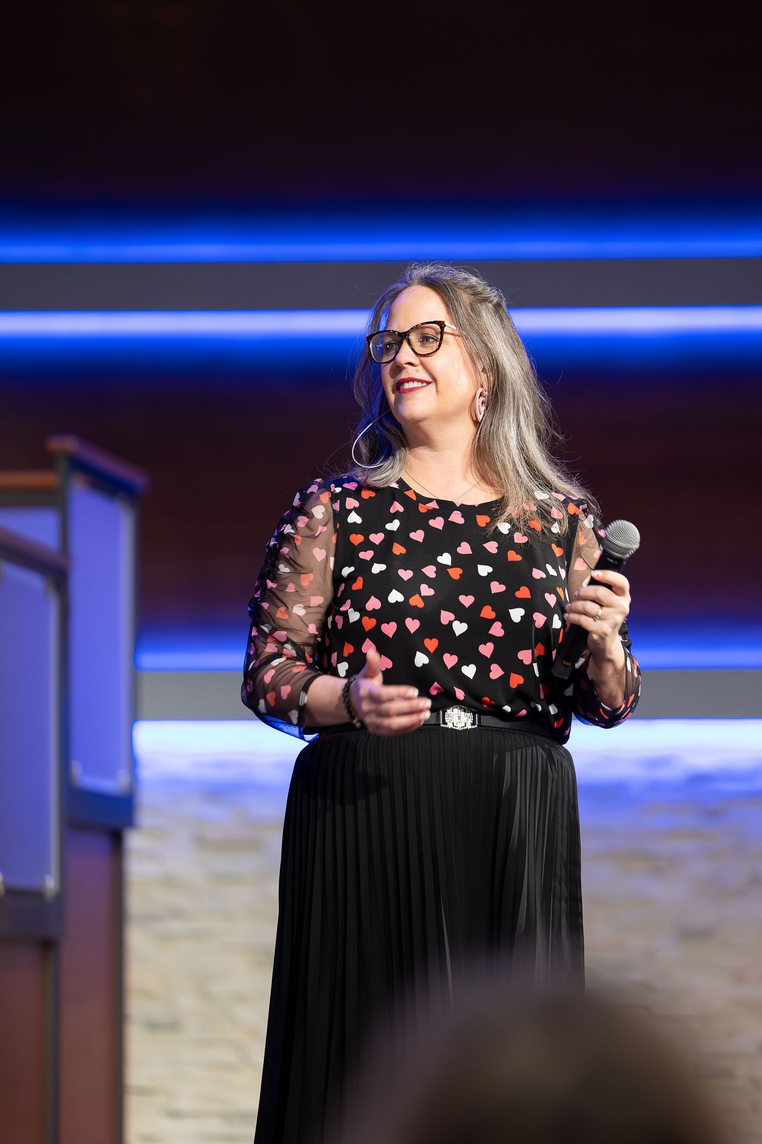Performer in heart-patterned dress with glasses sings on stage with blue lighting.