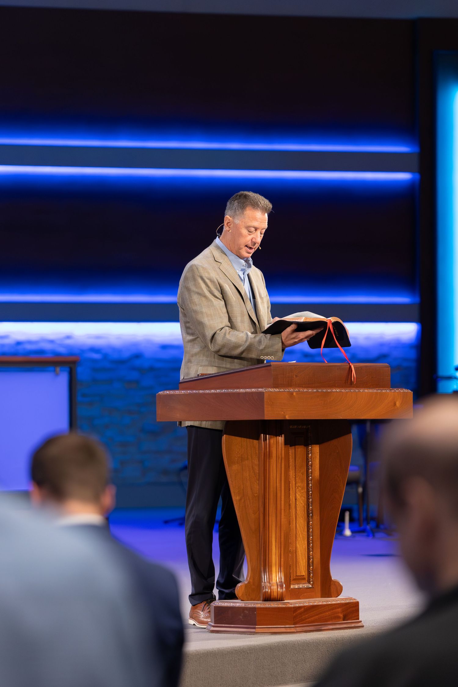 A speaker stands at a wooden podium on stage with blue accent lighting in the background.