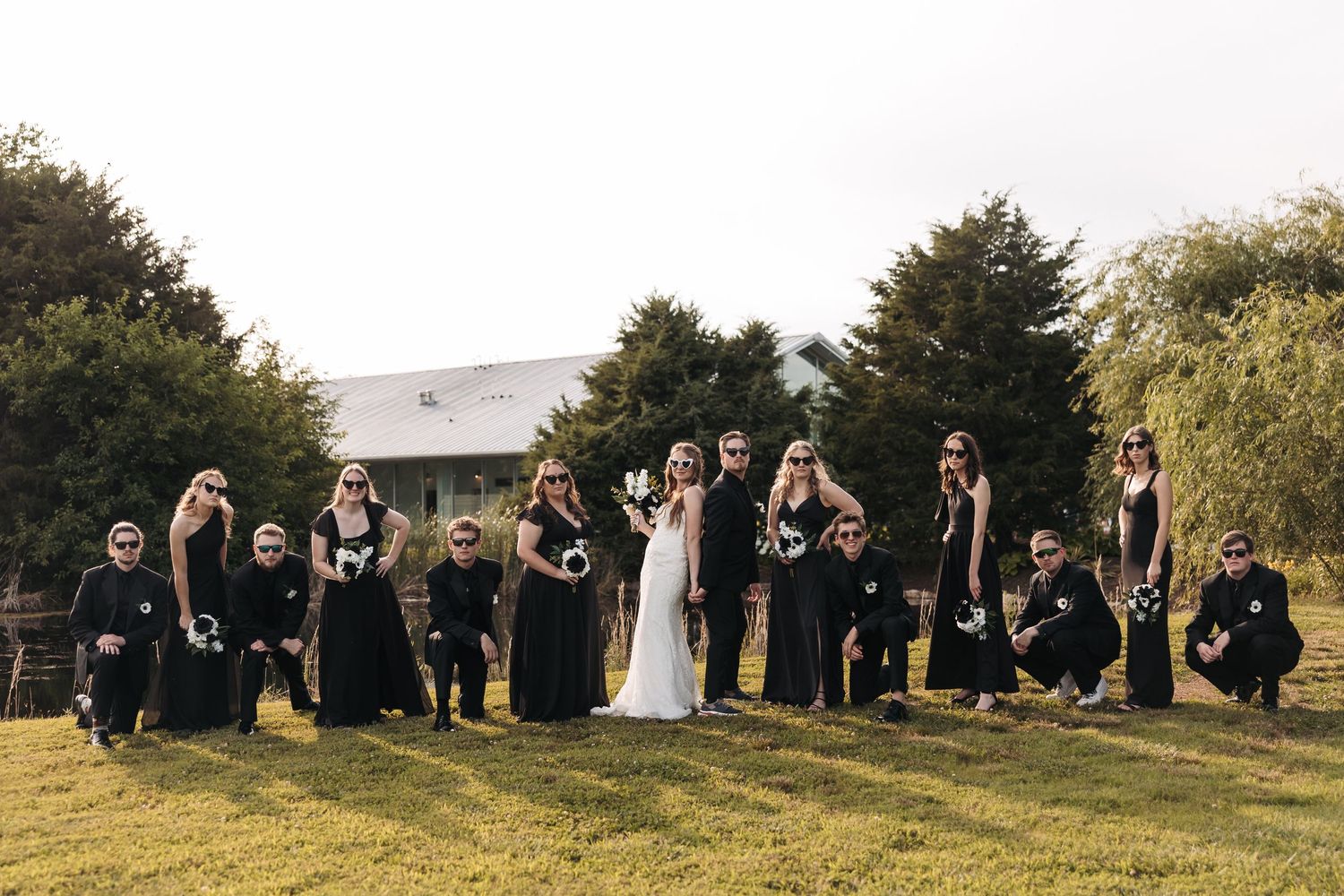Wedding party in black attire poses on grassy hill with trees in background during outdoor ceremony.