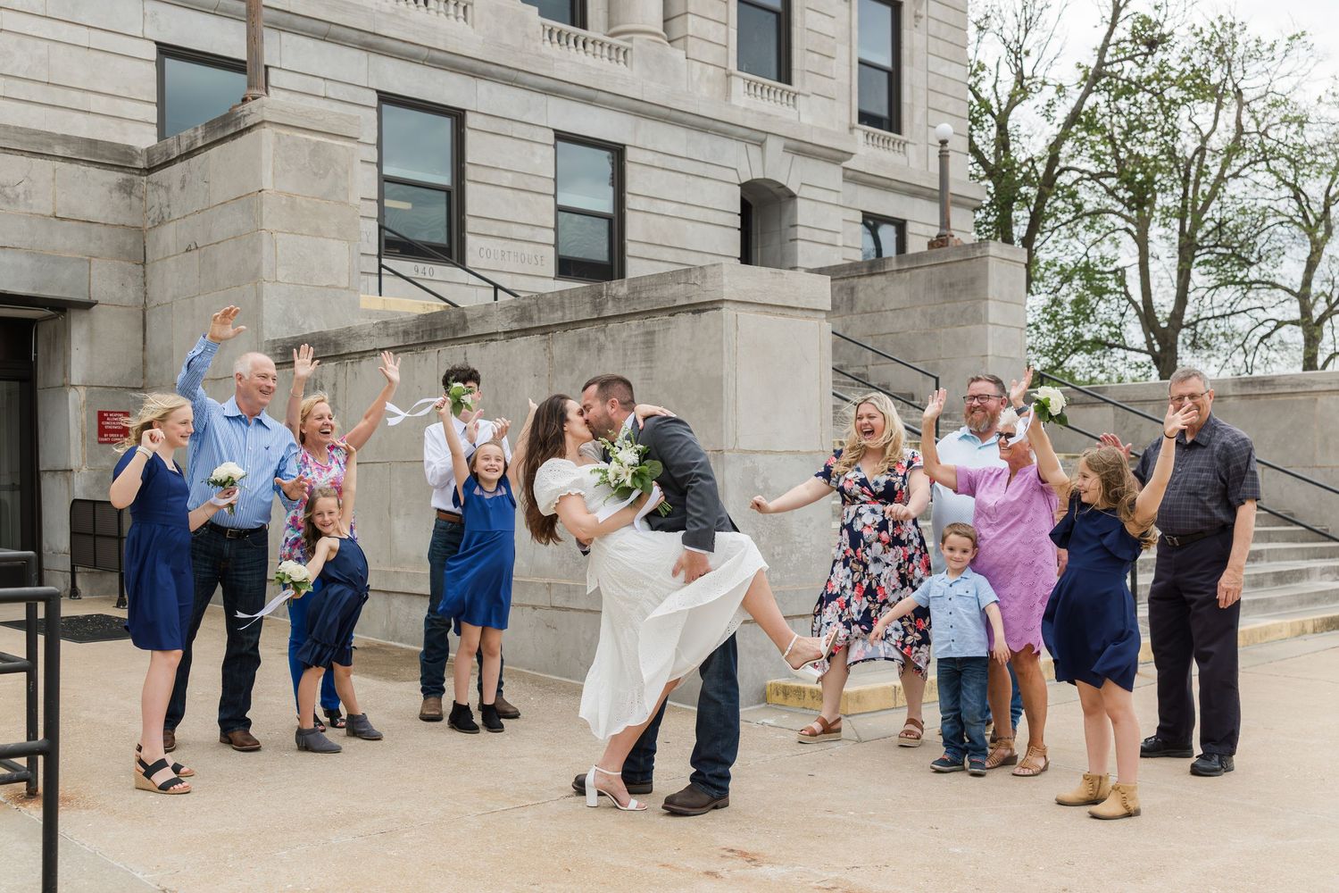 A couple shares a romantic dip while celebrating with a cheerful wedding party on stone steps outside a grand building.