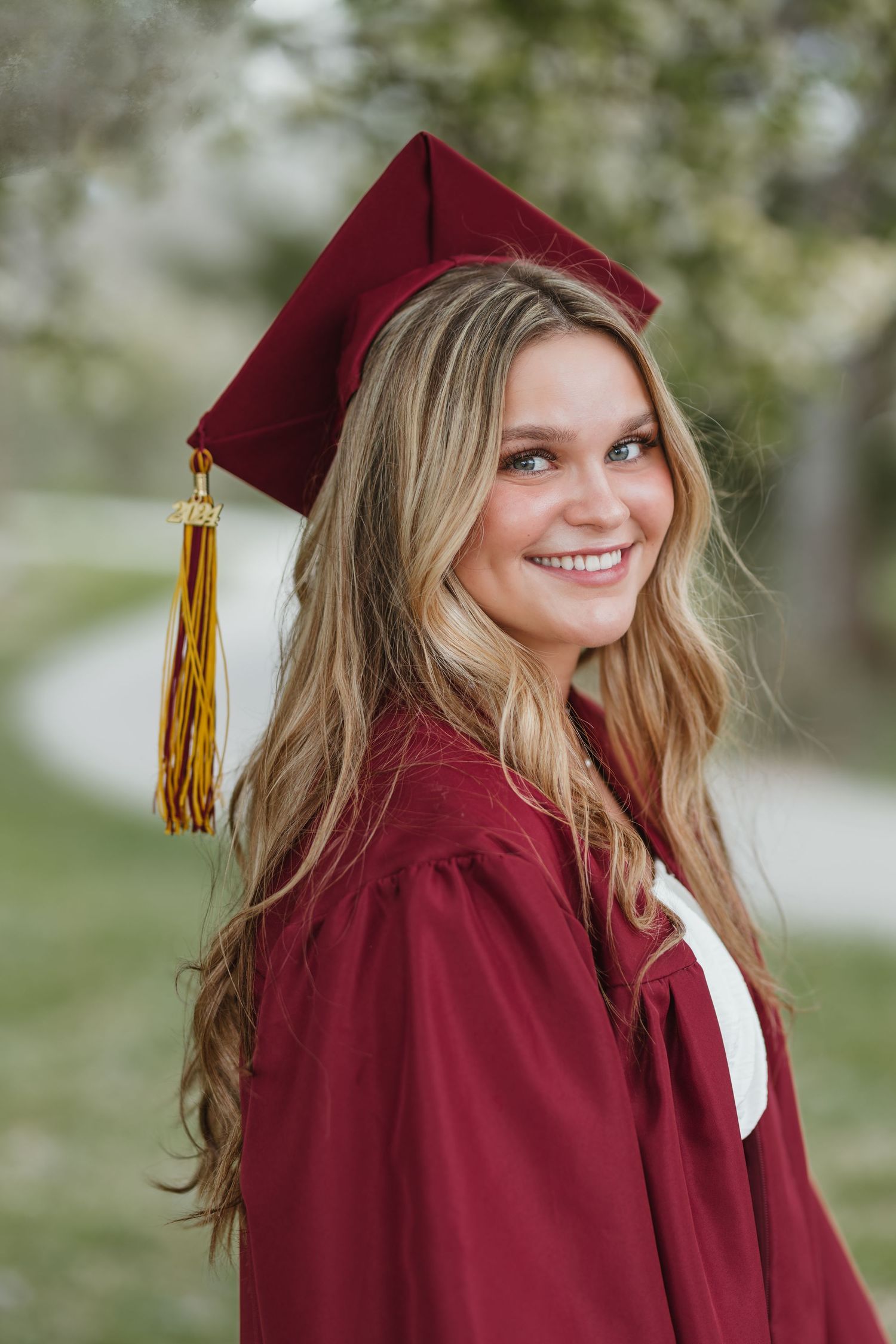A graduate in a burgundy cap and gown smiles while posing outdoors for a graduation portrait with a blurred background.