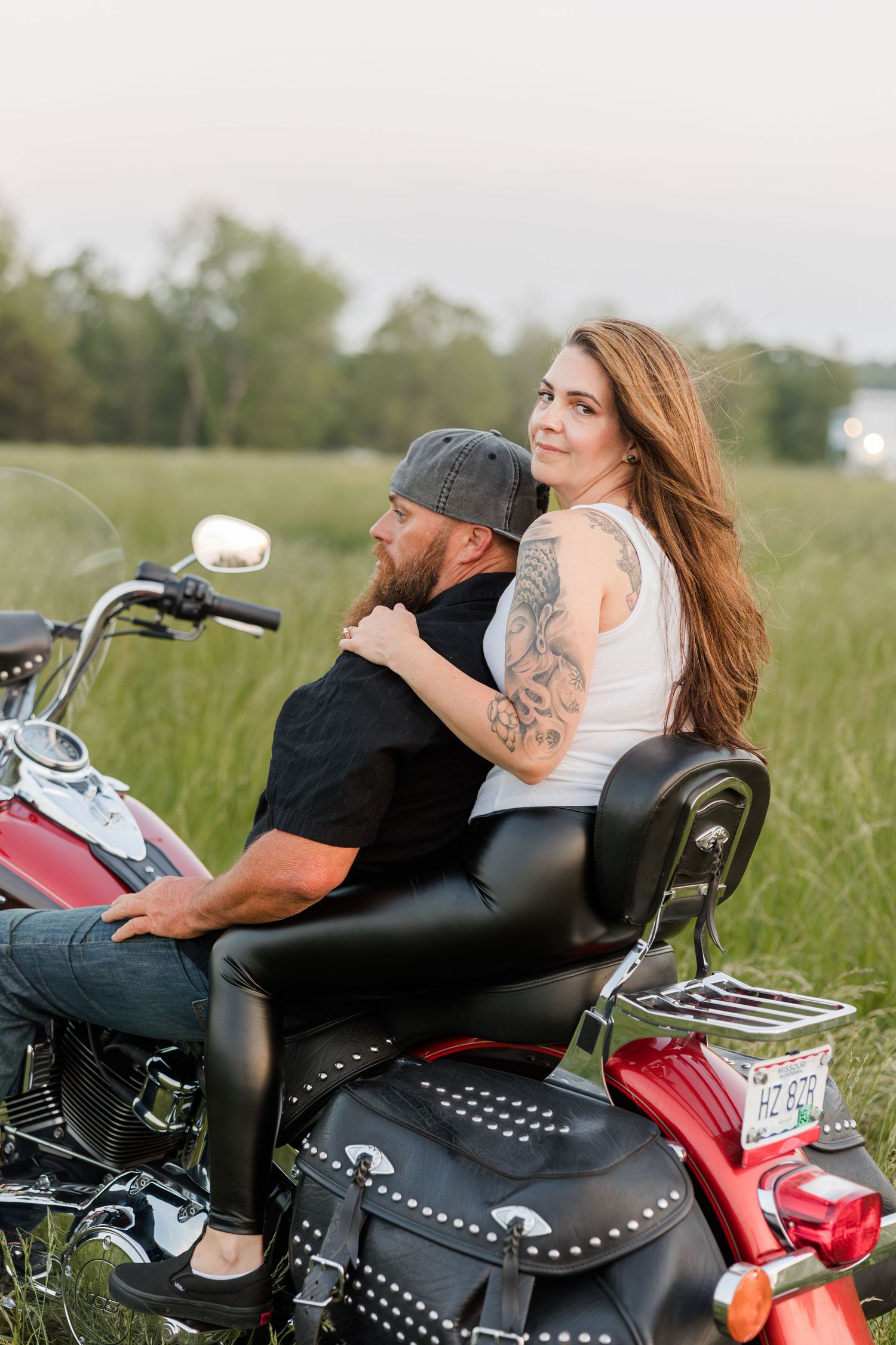 Two people seated on a red cruiser motorcycle with leather saddlebags in a grassy field at dusk.