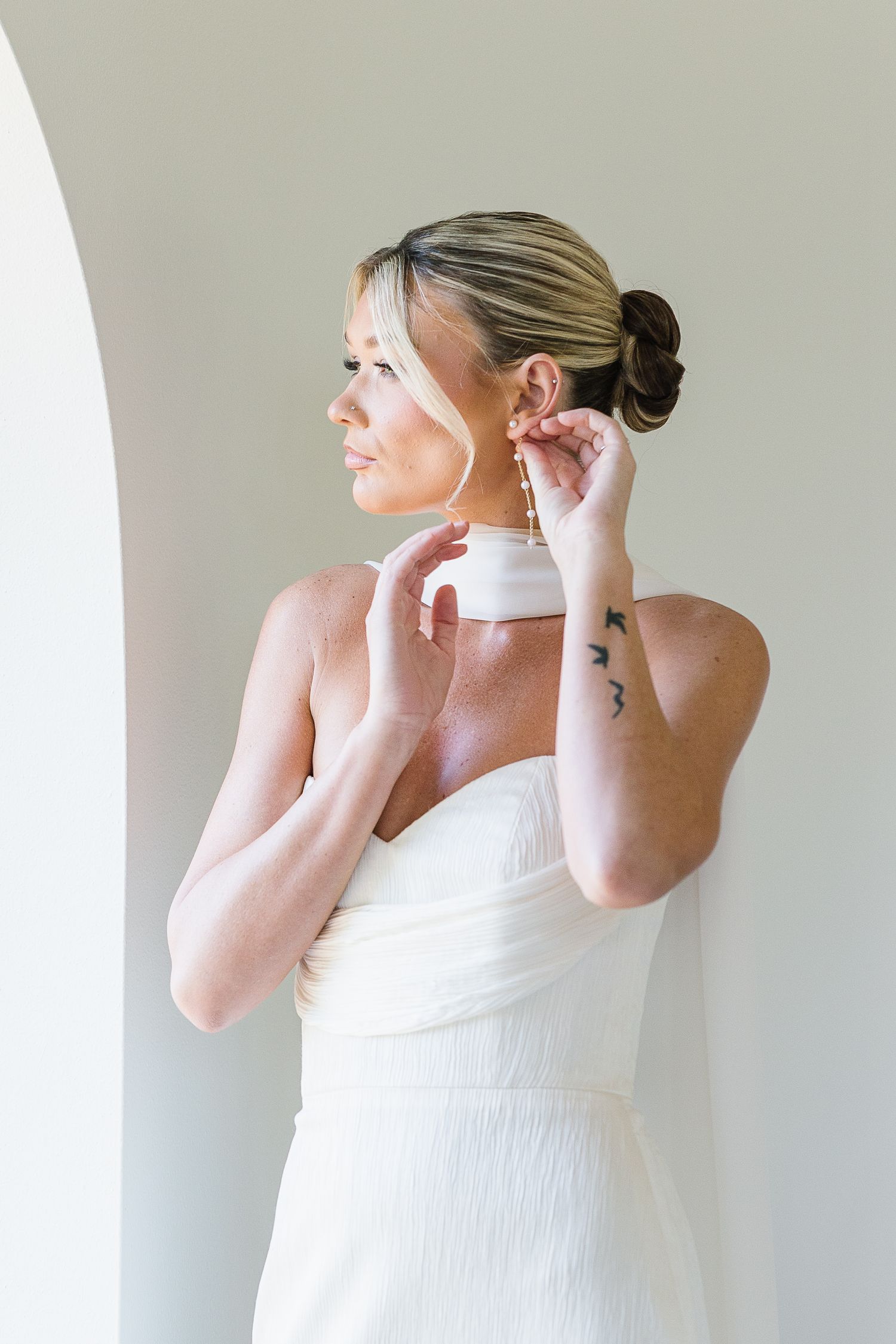 Woman wearing white tank top puts on earrings while getting ready against neutral background.