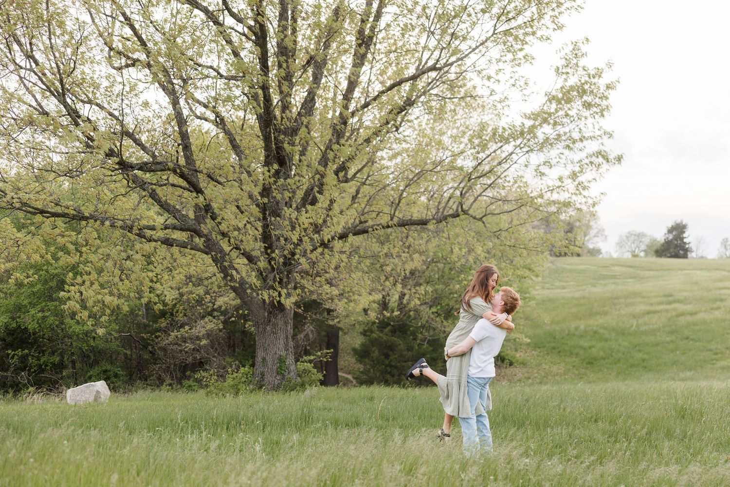 Two people embrace while dancing in a green grassy field near a large blooming tree in springtime.