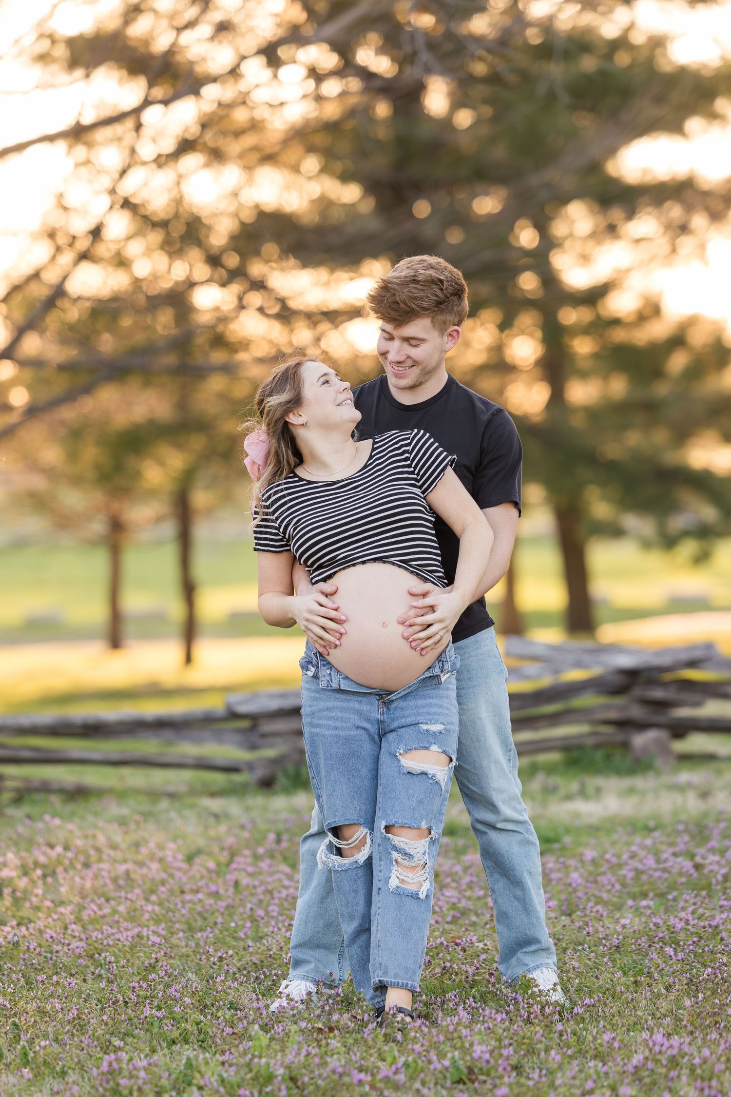 A couple shares an intimate moment together in a sunlit field with rustic wooden fencing in the background.