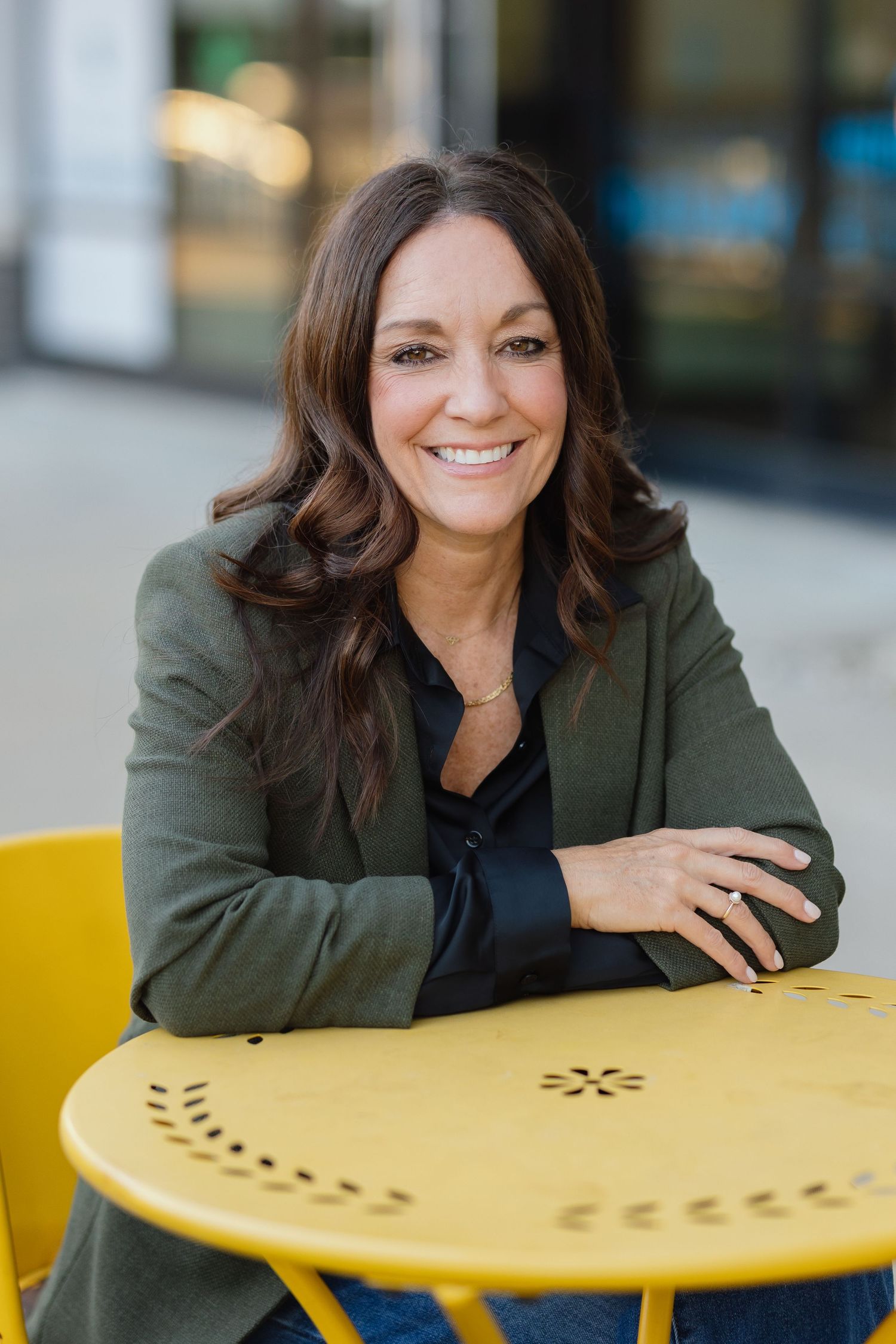 Professional headshot at yellow cafe table wearing olive blazer and black shirt.