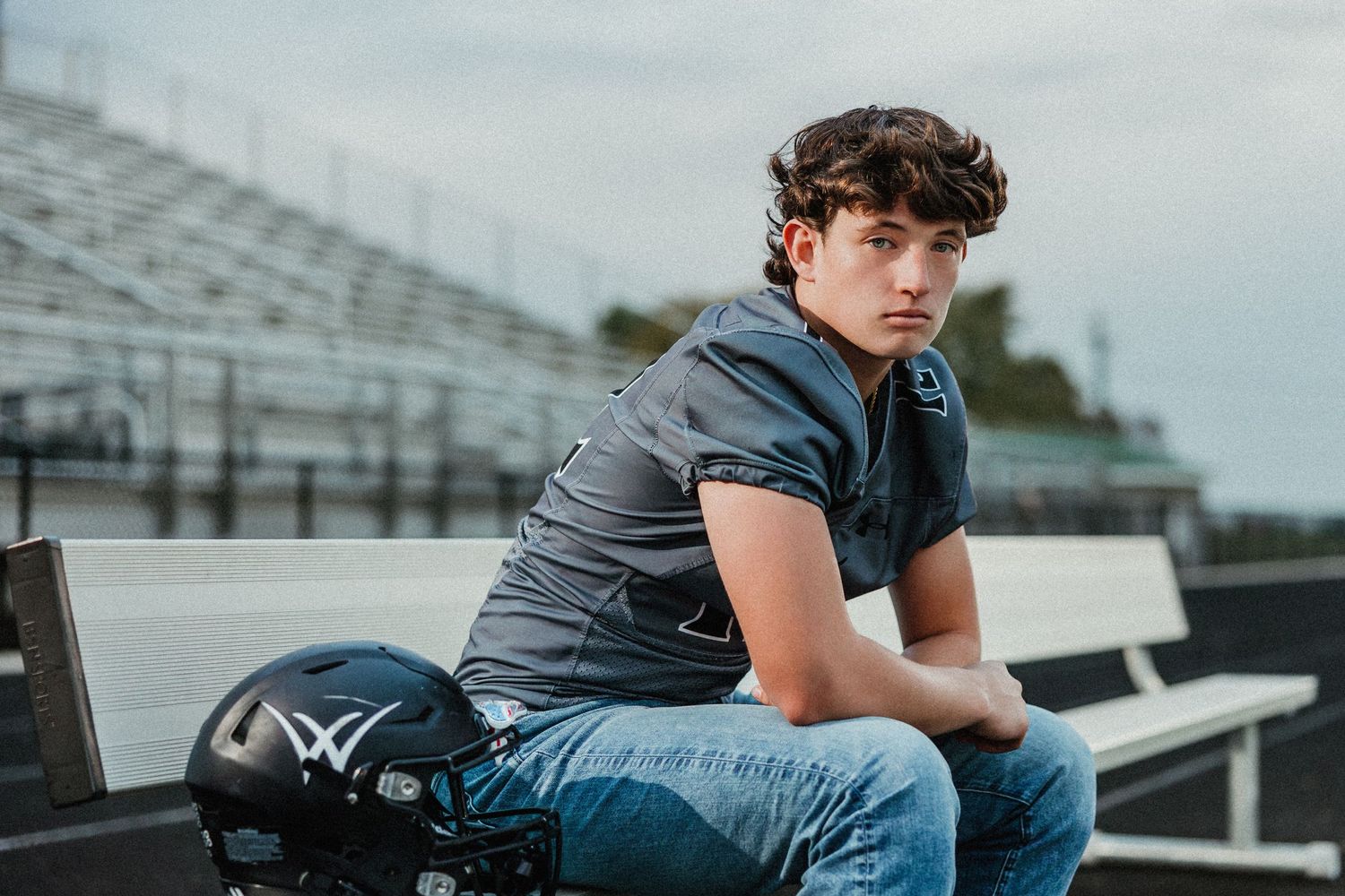 High school football player sits on stadium bleachers wearing gray jersey and holding helmet.