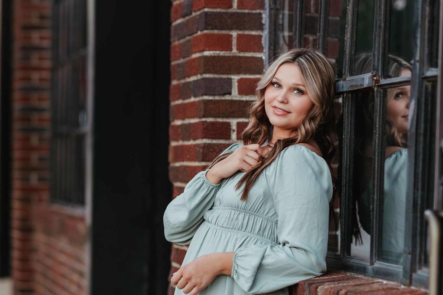 Person in sage green dress poses against brick wall in urban setting.