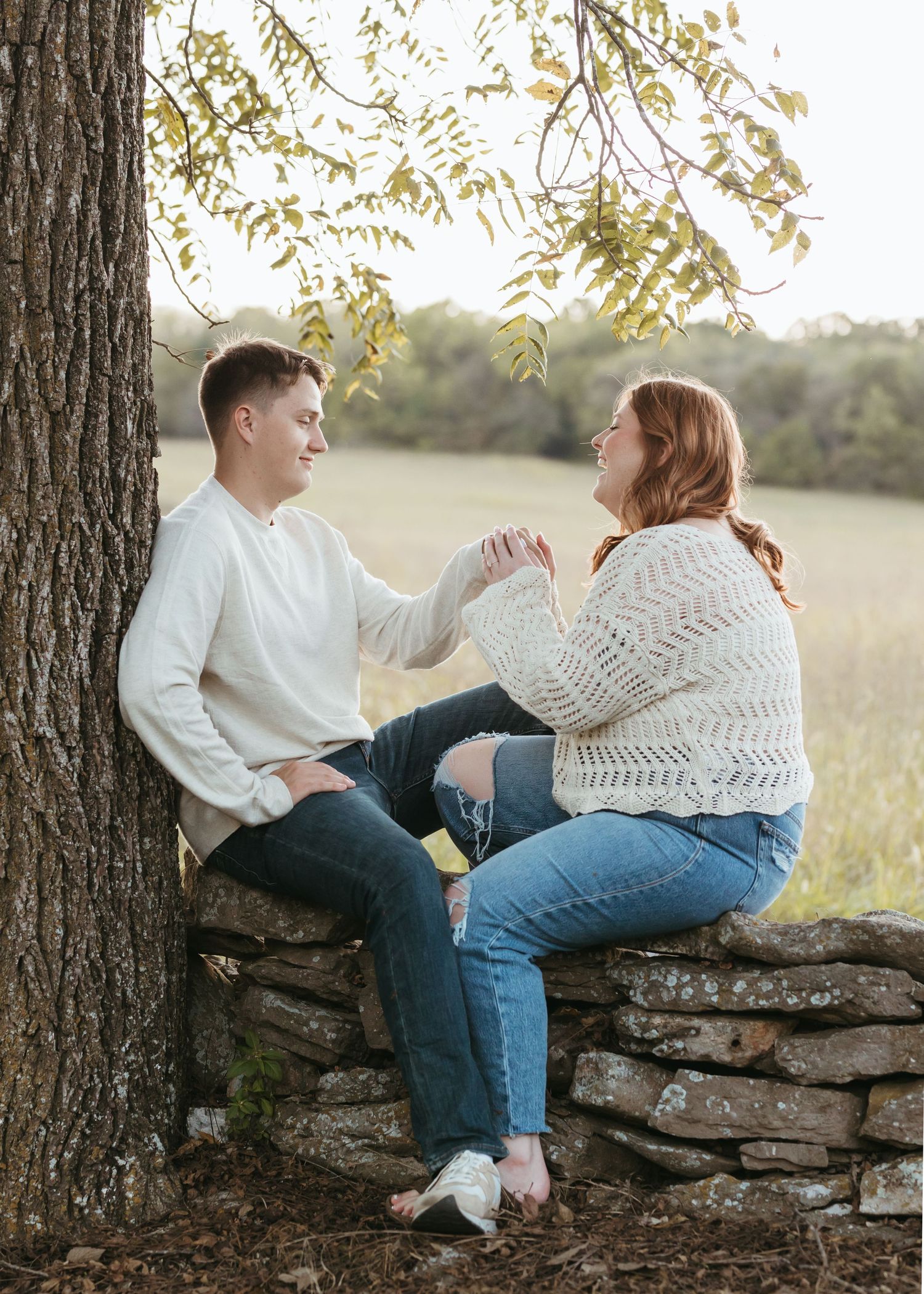 A couple sits on a stone wall beneath a tree in a golden field.