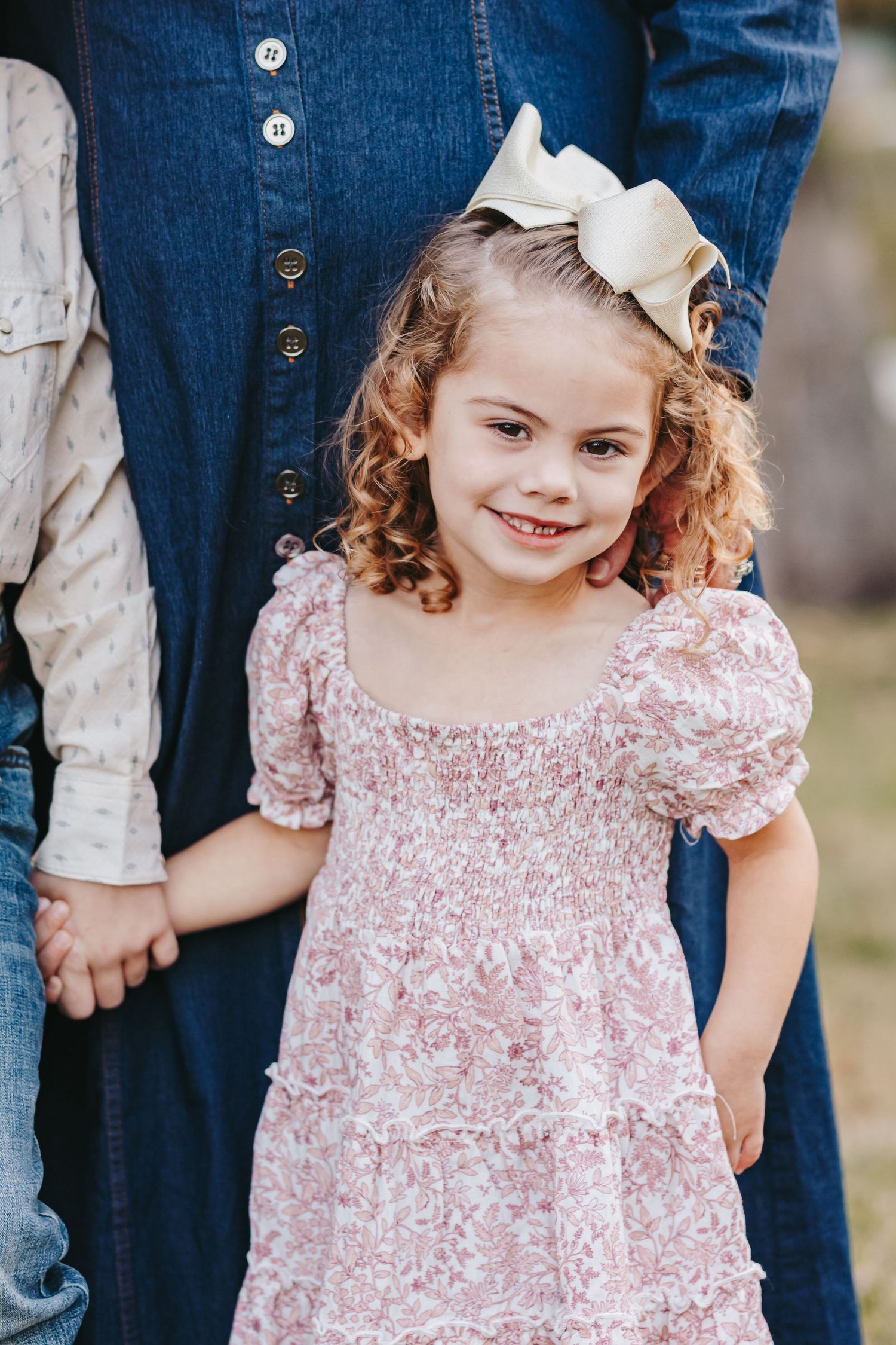 Young child in pink floral dress with bow in curly hair poses outdoors.