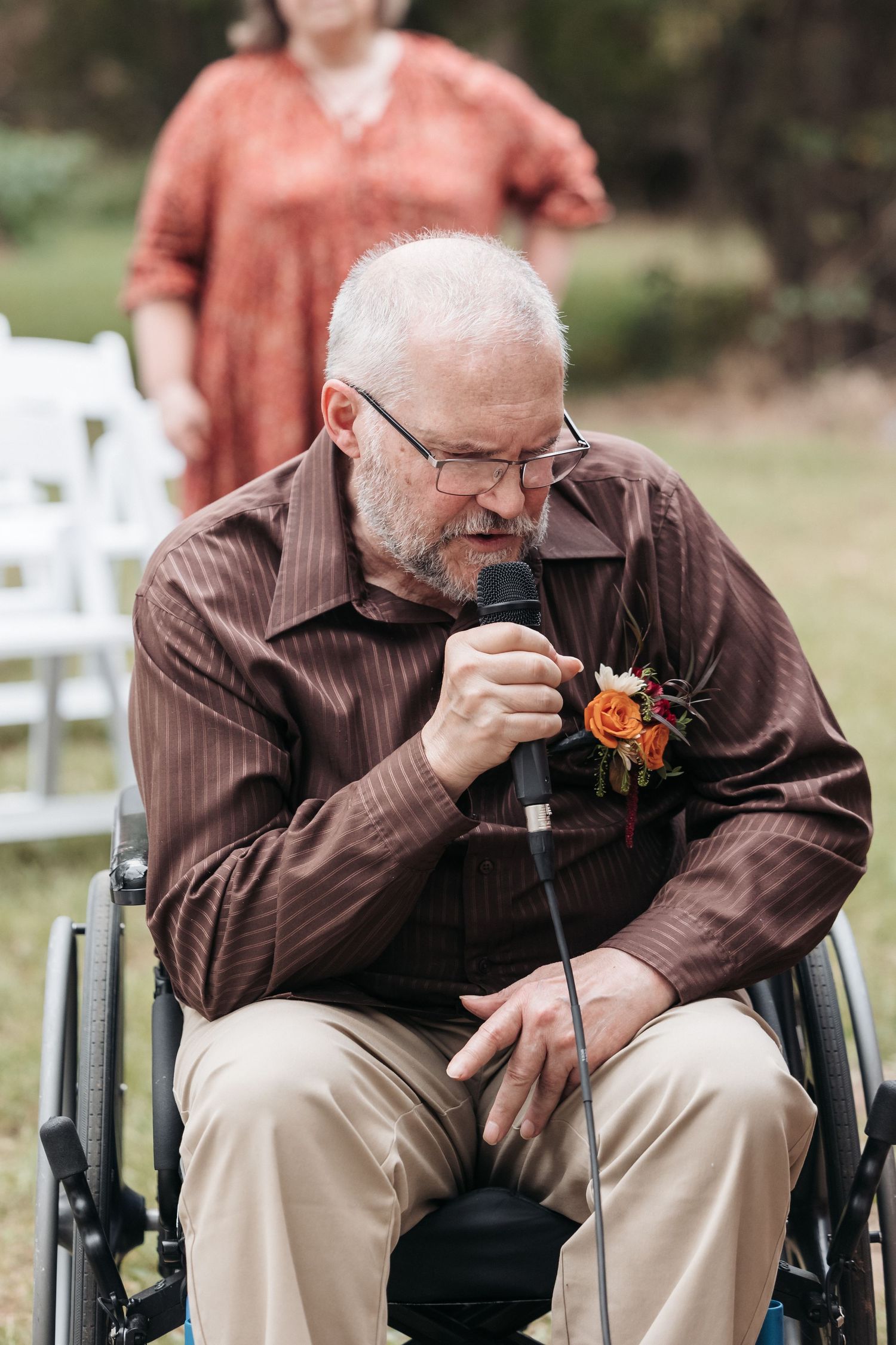 Person in a wheelchair wearing a brown striped shirt examines a coral-colored boutonniere at an outdoor event.