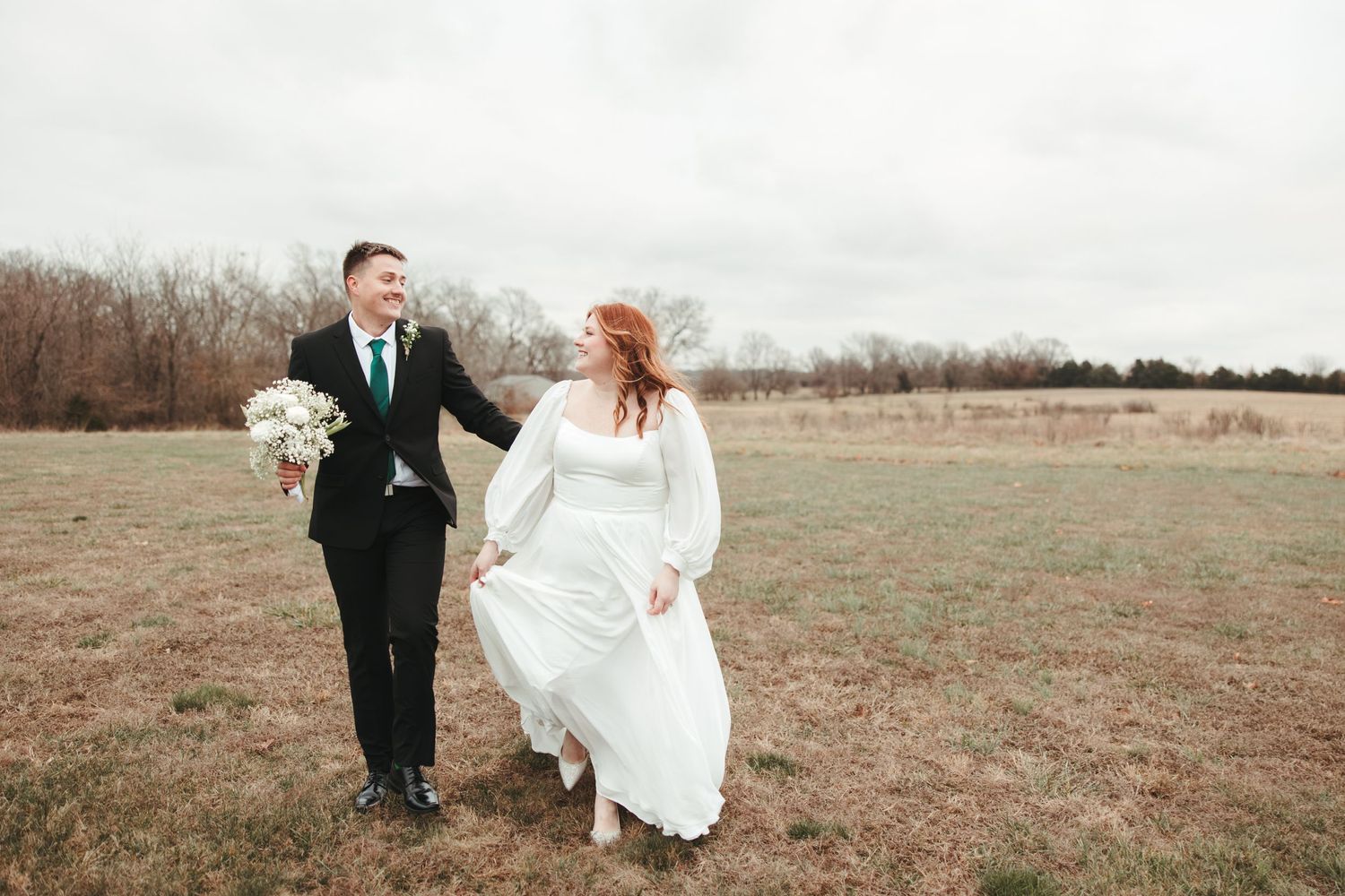A wedding couple walks together in a field during a winter wedding, with the bride wearing a flowing white dress.