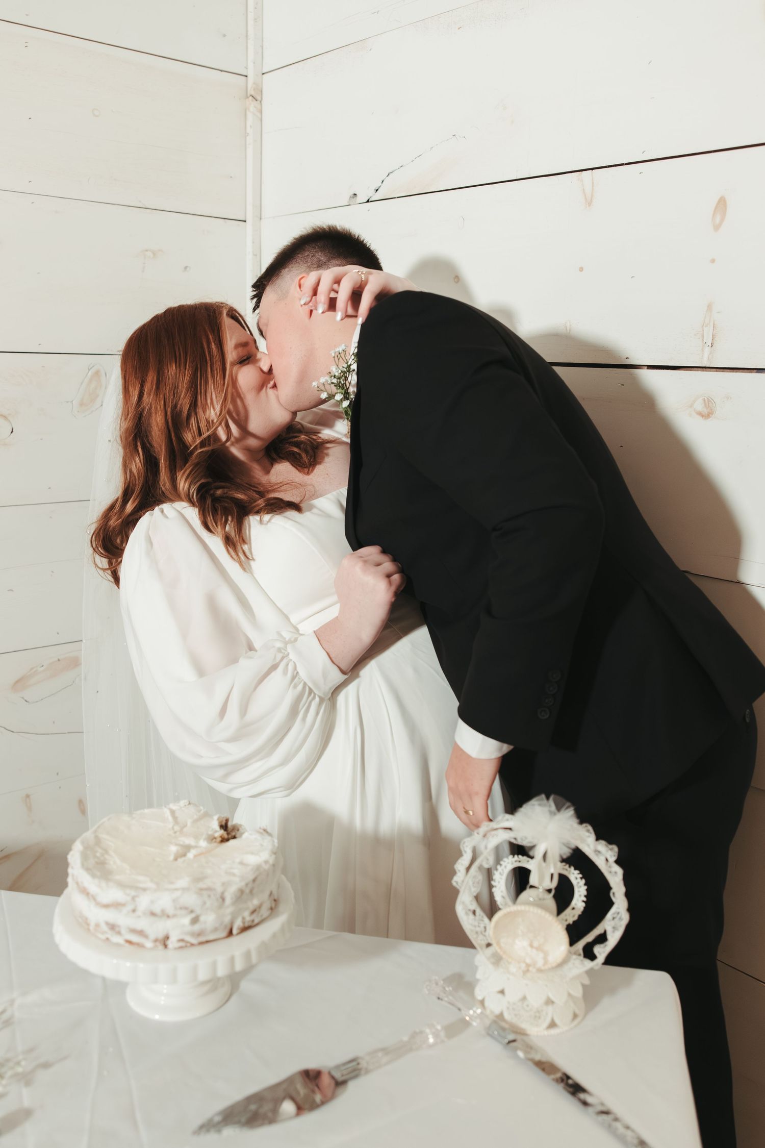 Newlyweds share a playful moment while cutting their wedding cake against a white wall.