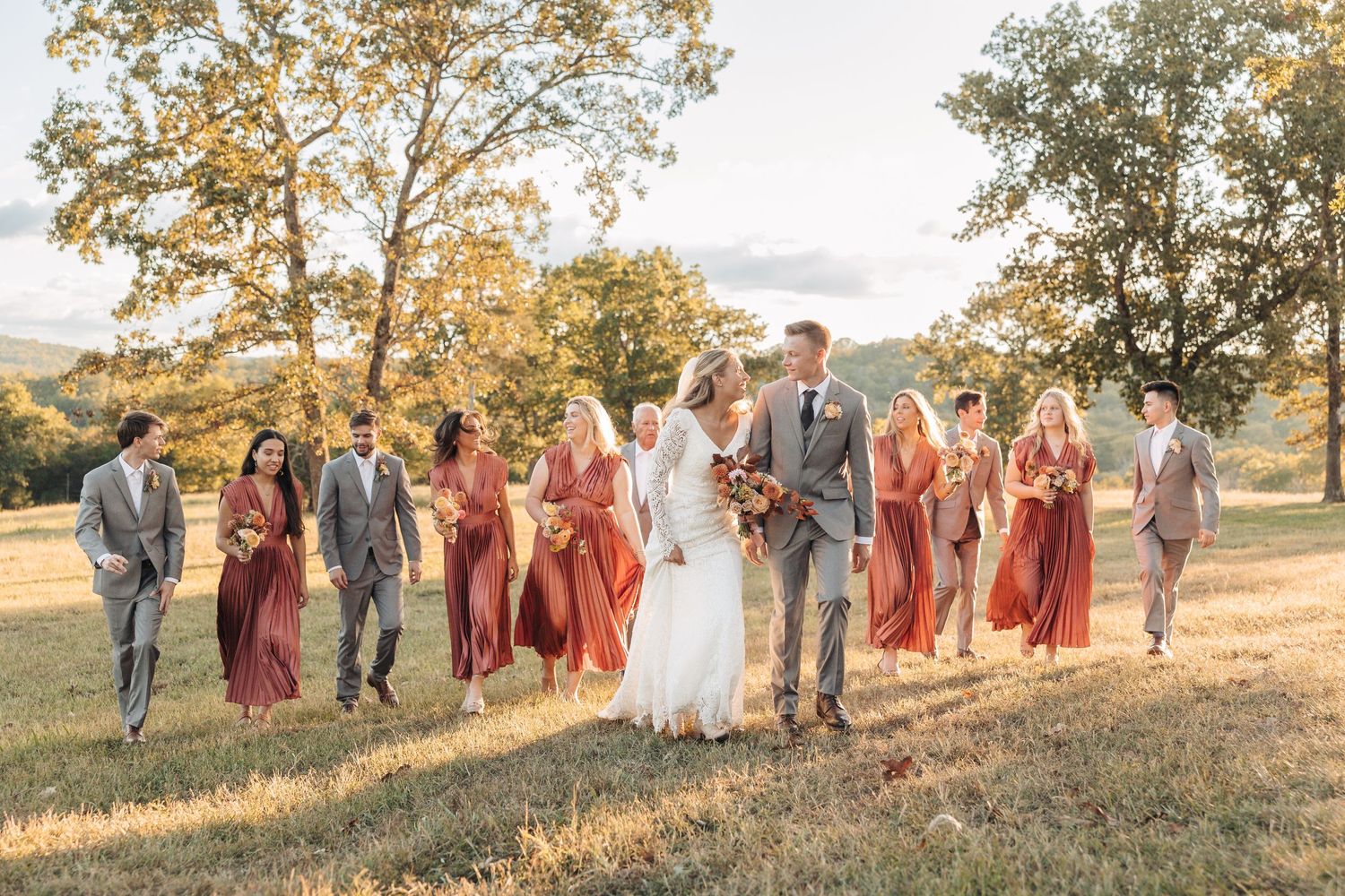 Wedding party walking together on grassy hill wearing burgundy and gray attire at sunset.