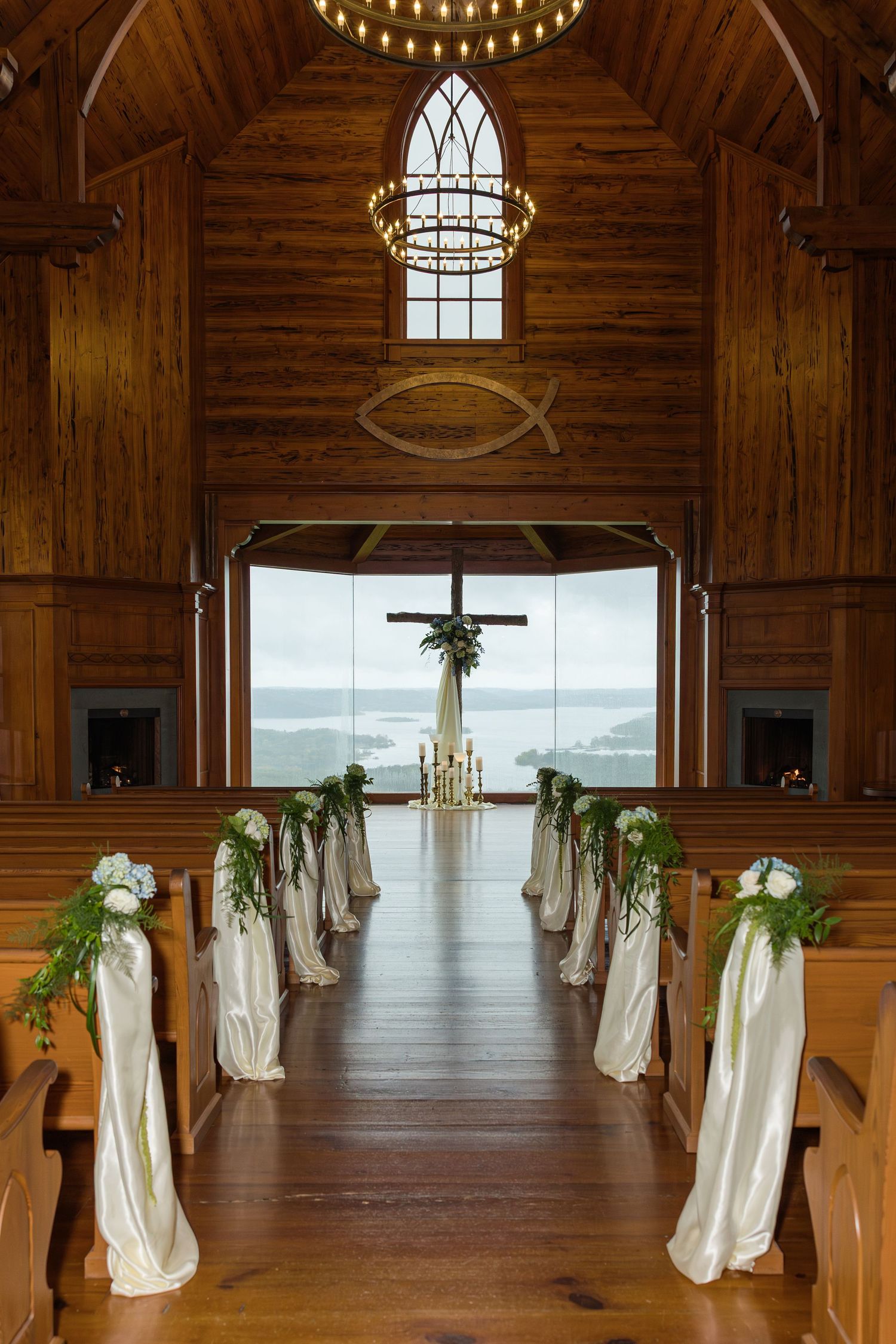 Rustic wooden church interior with white fabric draped pews and chandeliers leading to a large window overlooking water.