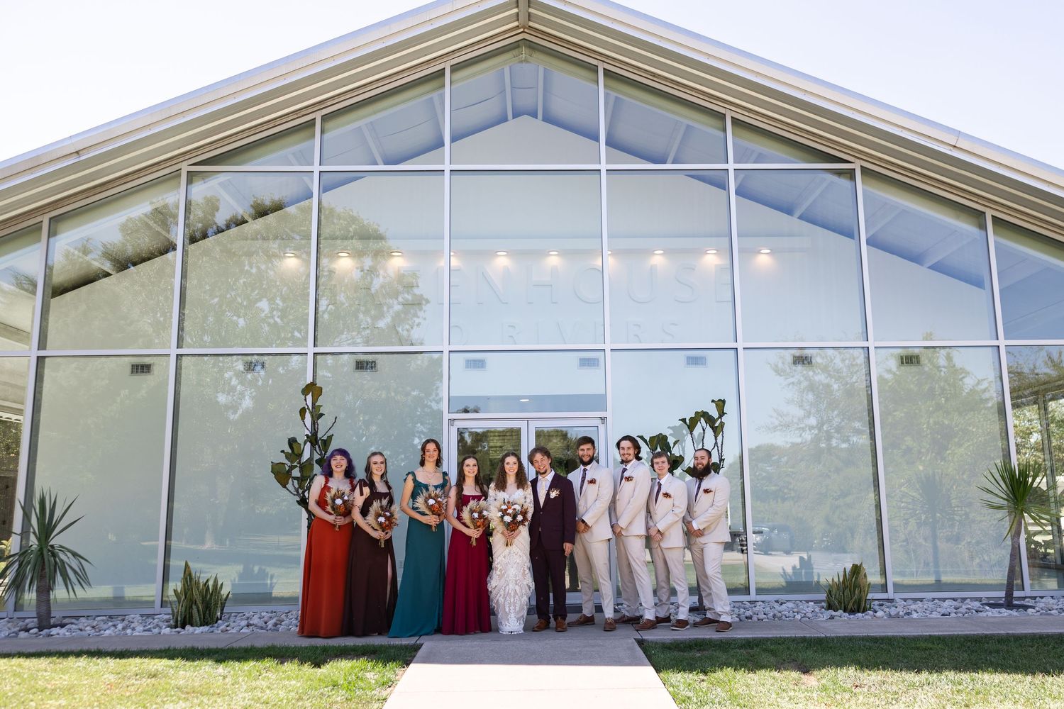 Wedding party poses in colorful dresses and tan suits outside a modern glass venue building.