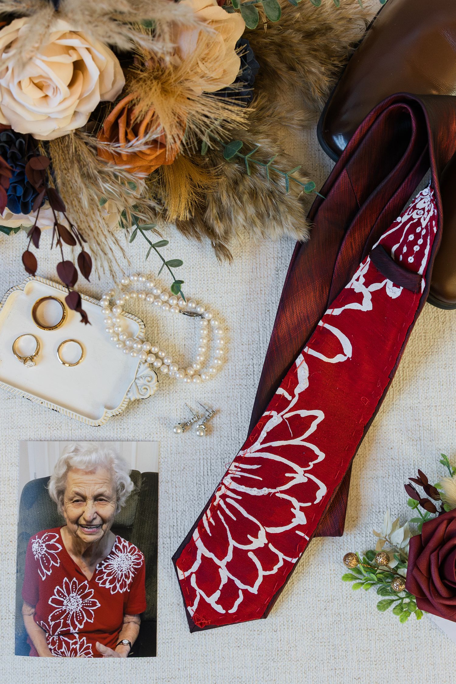 Wedding flat lay with red floral tie, pearl jewelry, photo of elderly woman, bouquet, and gold rings on linen.