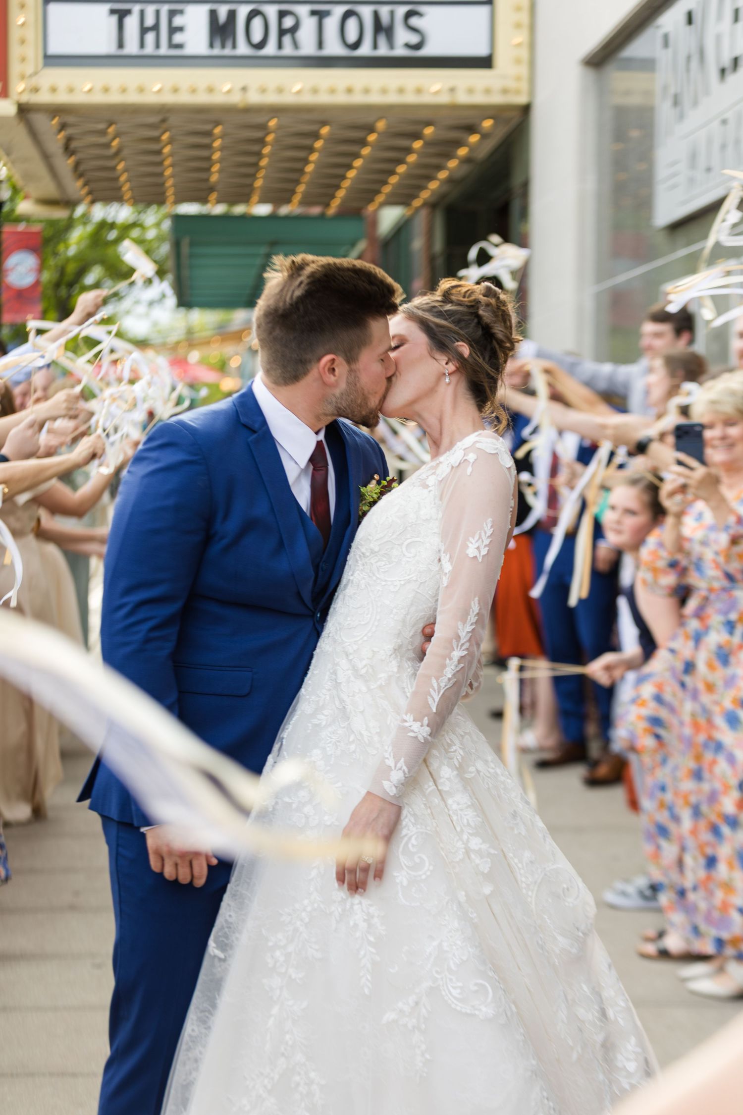 Bride and groom share a romantic kiss outside The Mortons theater marquee surrounded by cheering guests.
