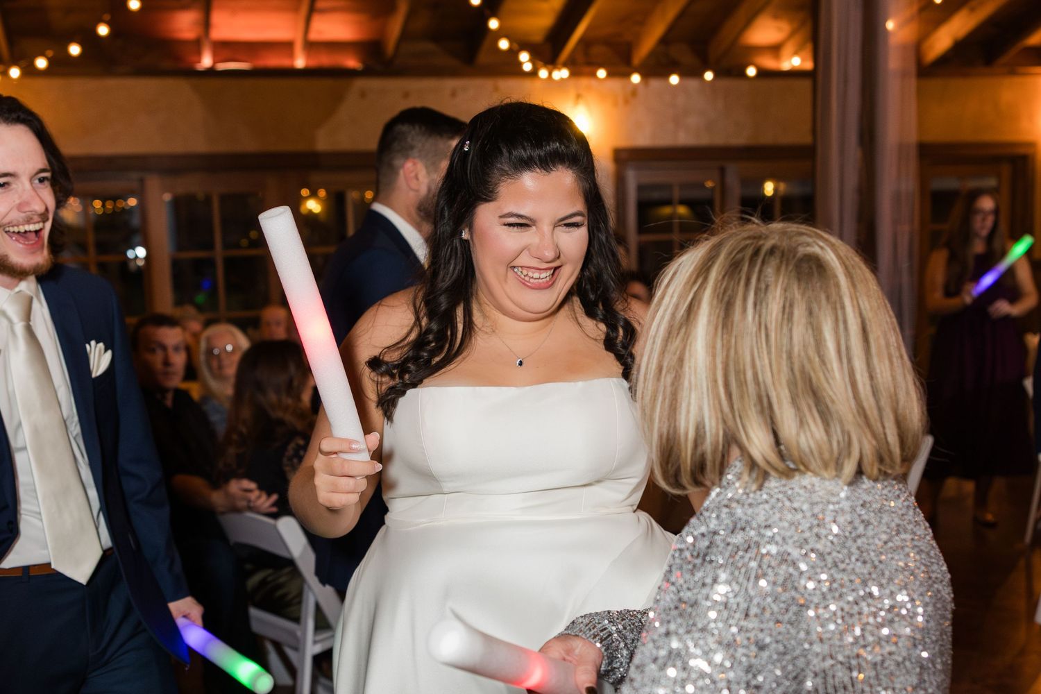 Bride in white gown holding glow stick at wedding reception, smiling and dancing with guests under warm string lights.