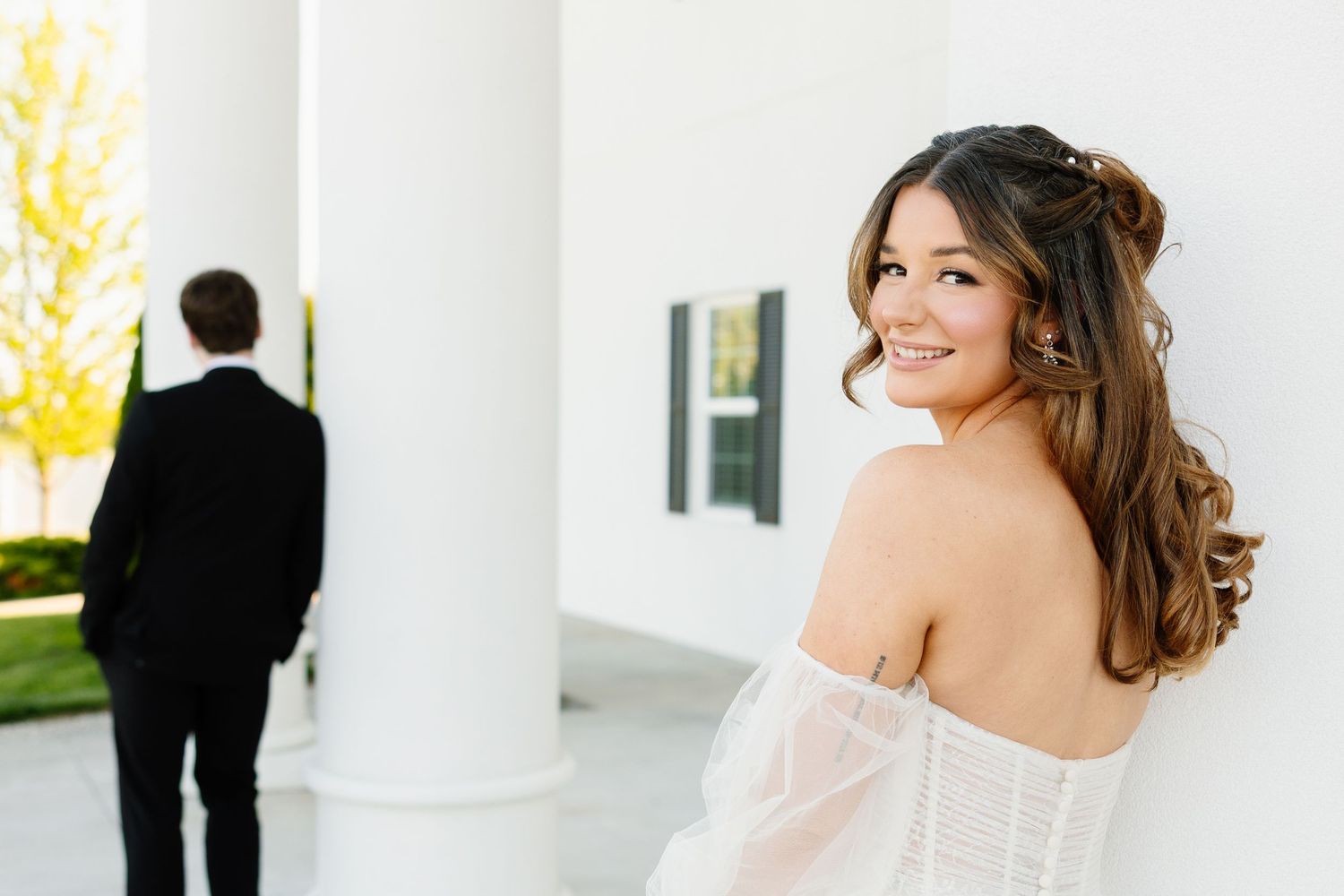 Bride in off-shoulder gown smiles over shoulder while groom stands by white columns outdoors.
