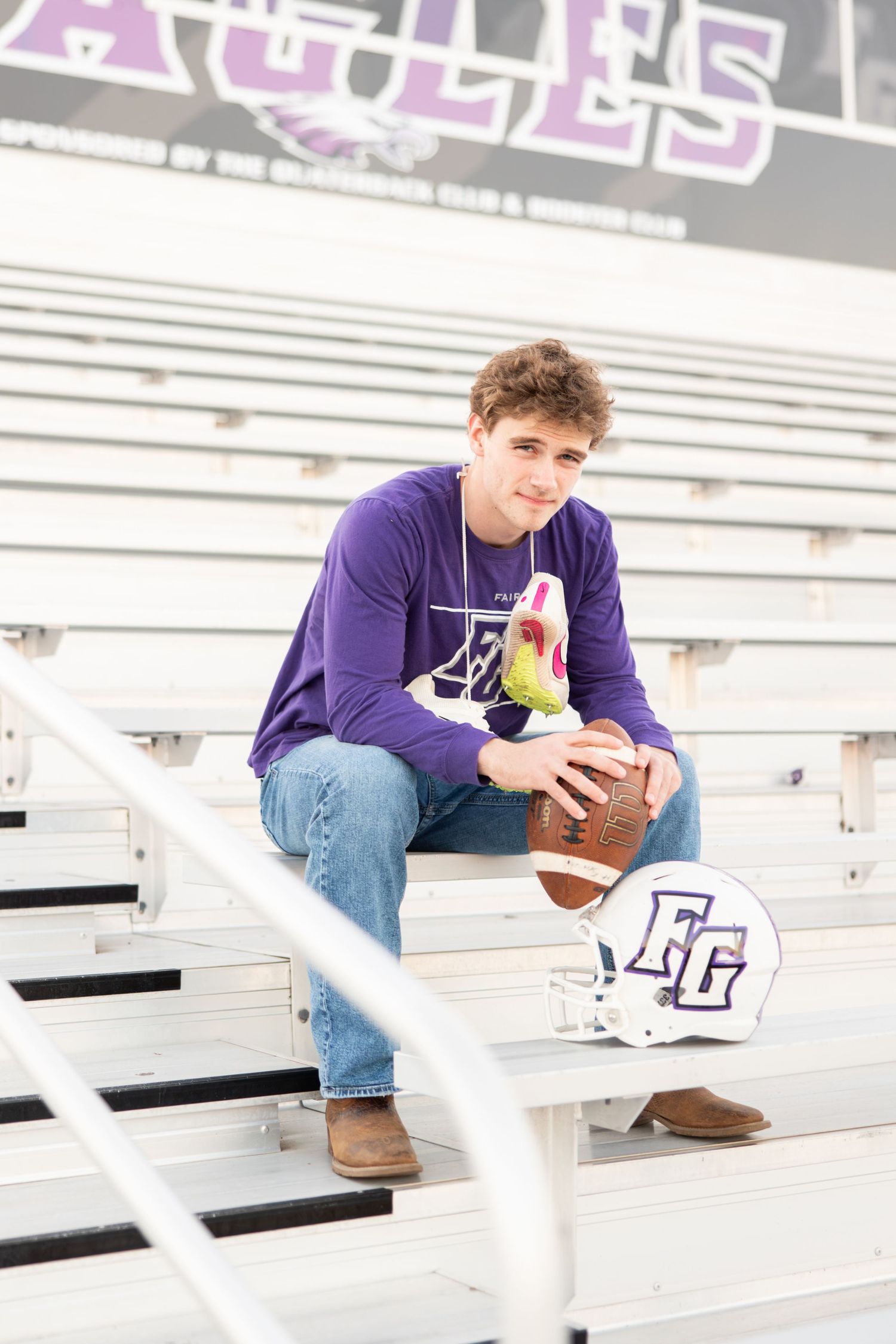 Person in purple hoodie poses with football on stadium bleachers next to team helmet.
