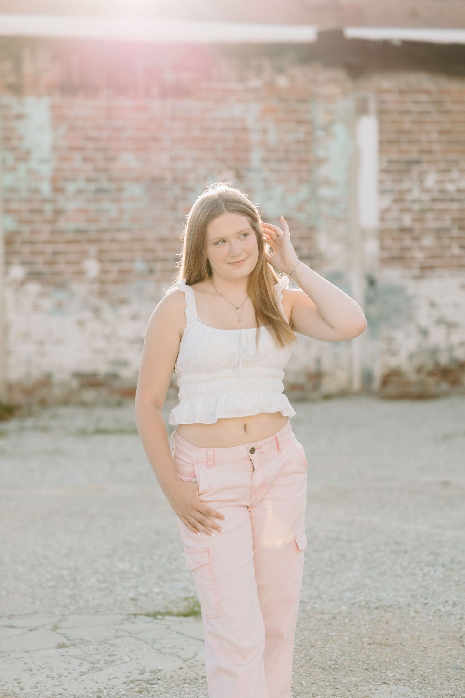 A casual fashion photoshoot outdoors features pastel pink pants and white crop top against a weathered brick wall.