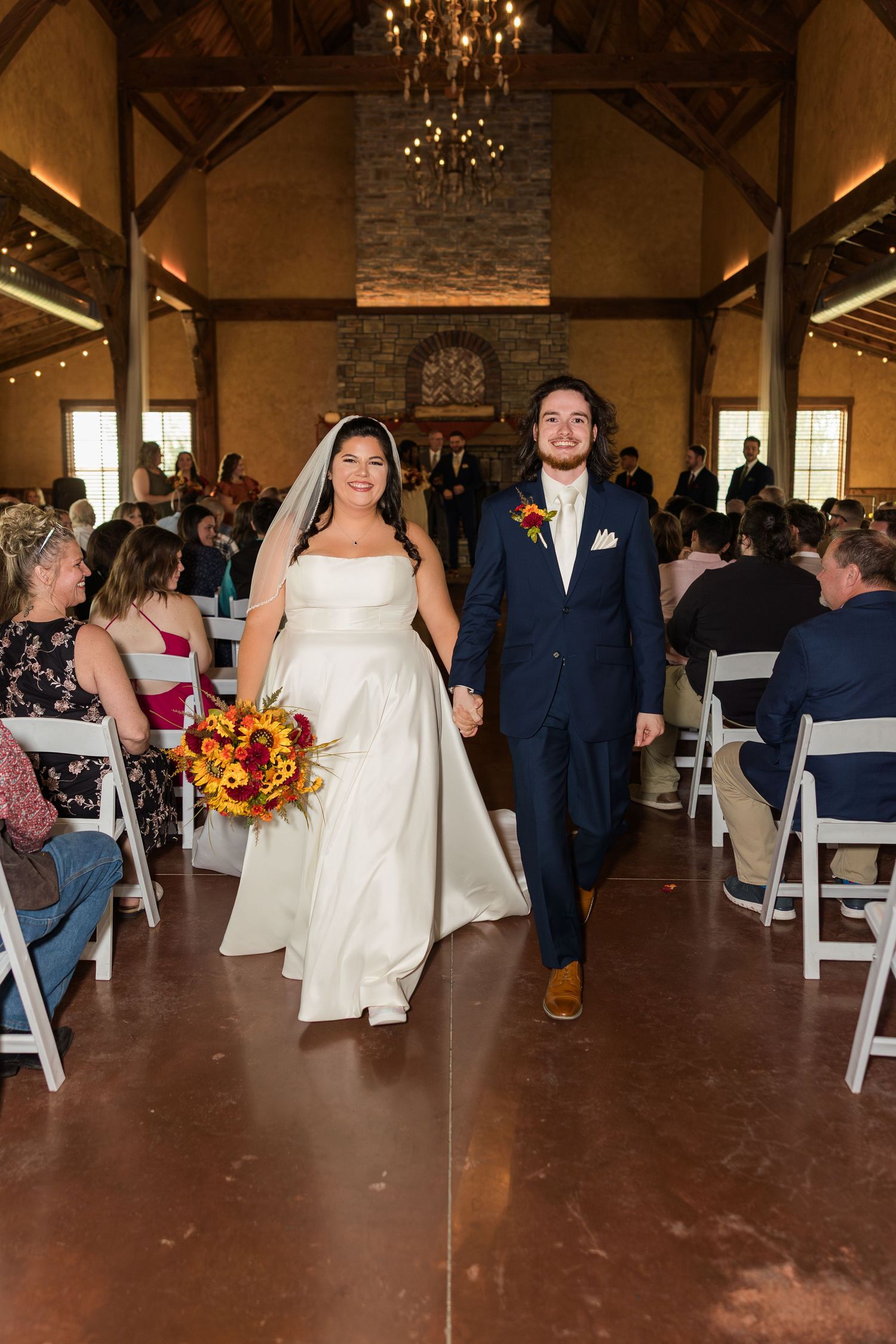Wedding ceremony in a rustic indoor venue with guests seated in white chairs and autumn decor.