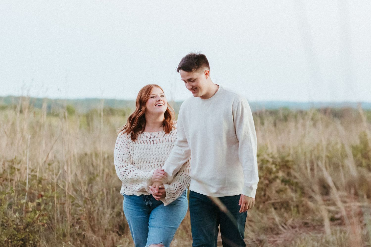 A couple walks hand in hand through tall grass in a scenic outdoor setting.